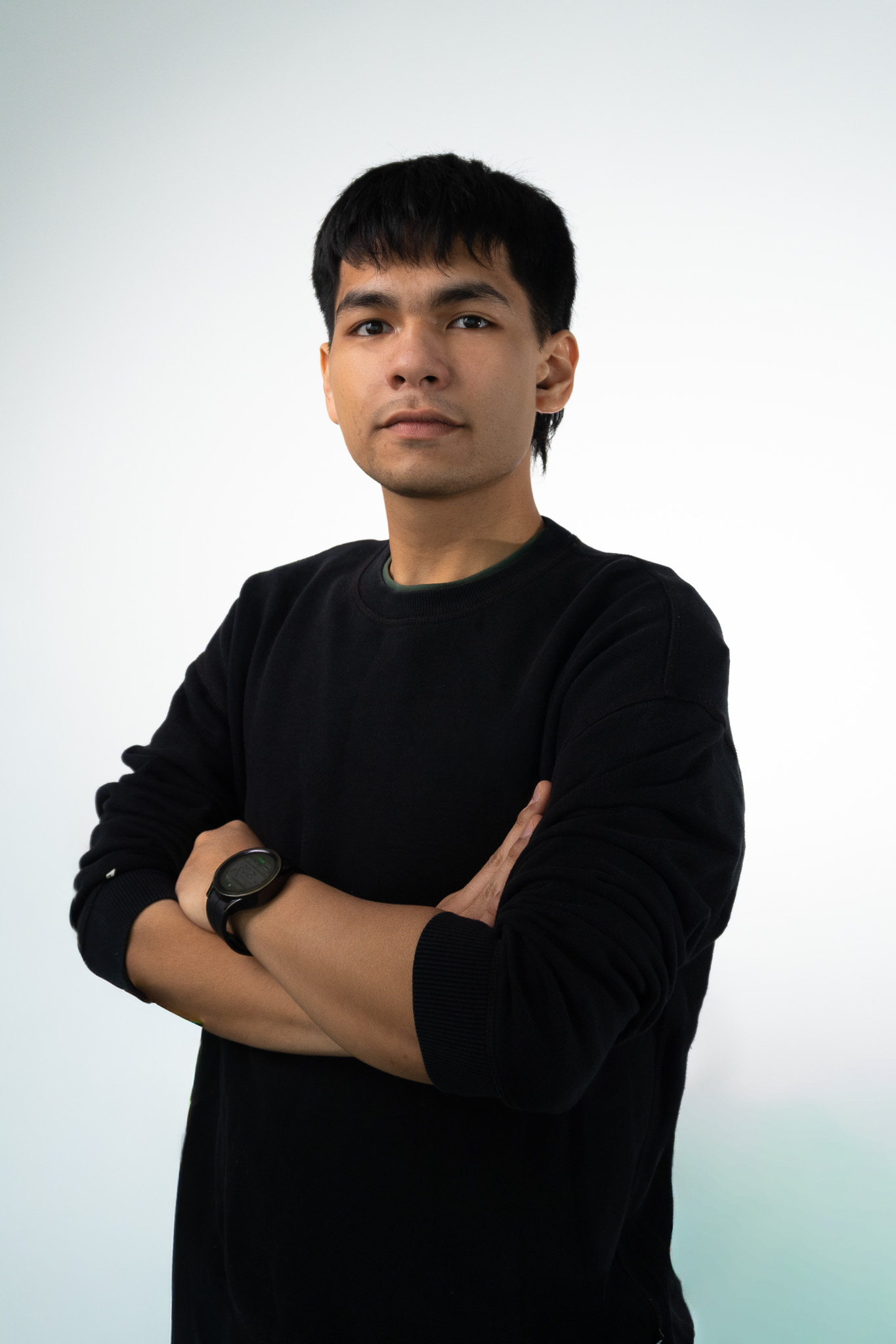 A young man with short dark hair wearing a black sweatshirt and a wristwatch, standing with arms crossed against a plain white background.