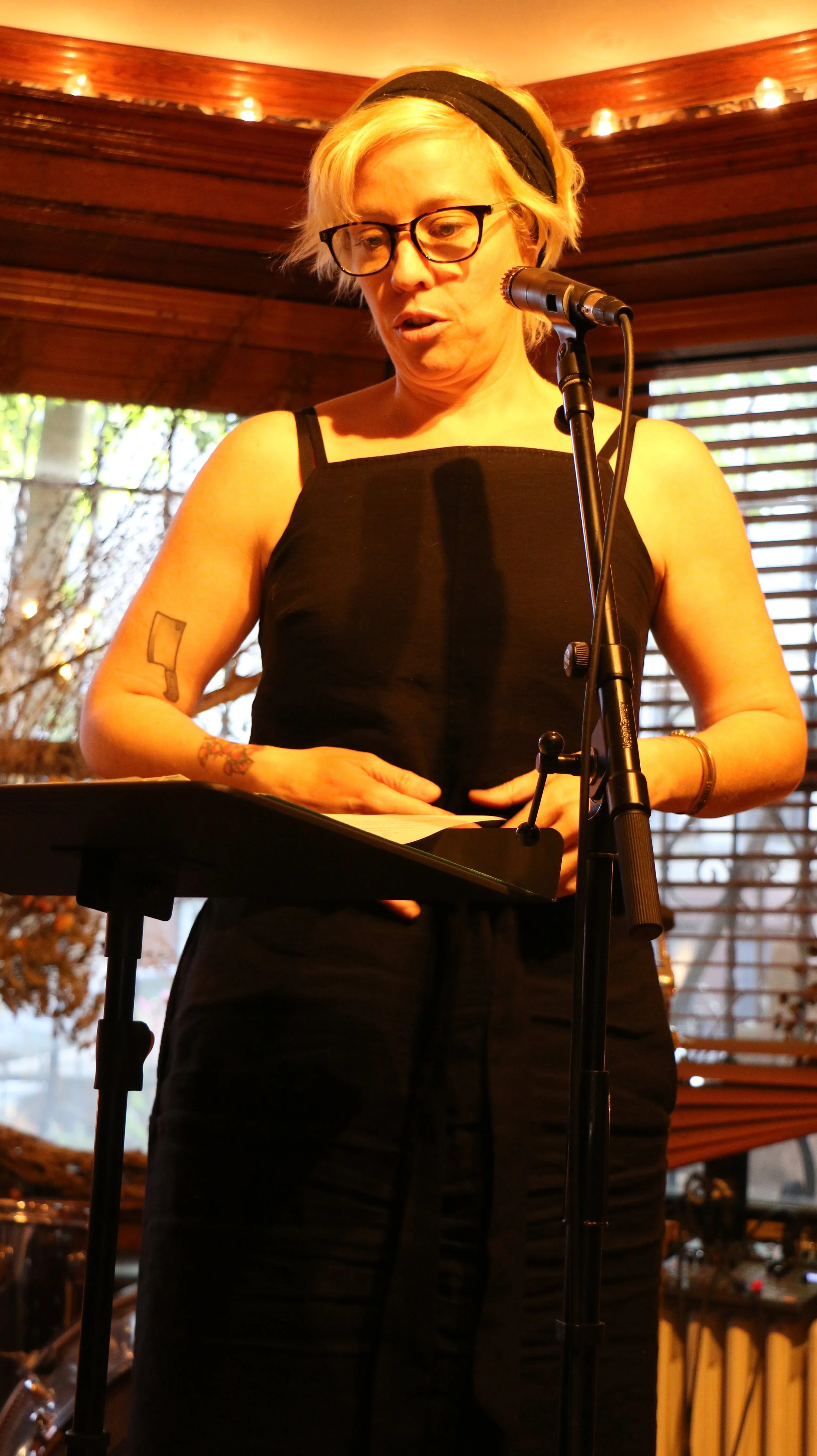 Person wearing glasses and a black dress speaking into a microphone at an indoor event.