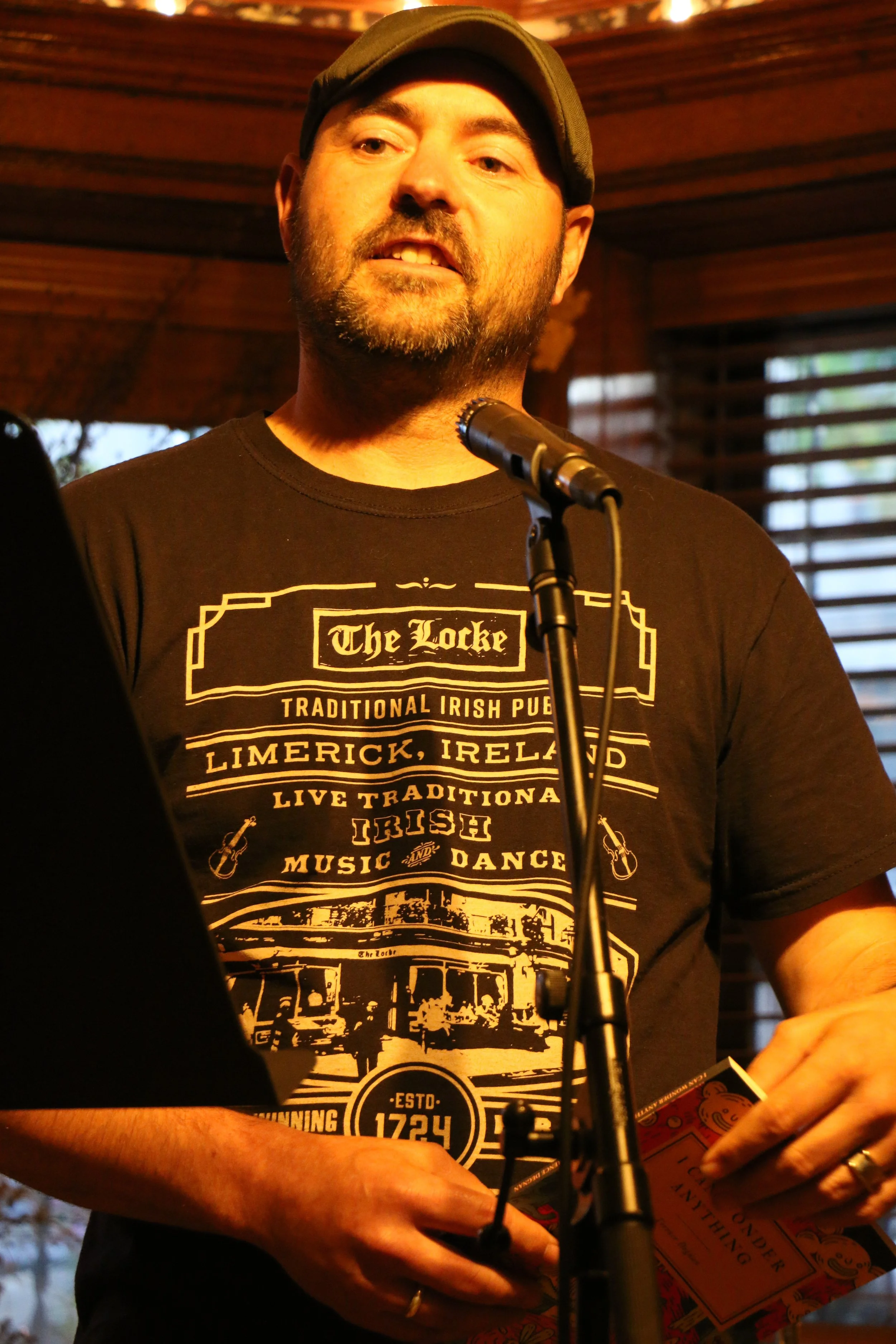 Man wearing a black "The Locke" t-shirt, speaking into a microphone, holding a book. The shirt mentions "Traditional Irish Pub" in Limerick, Ireland, with graphics of instruments and a pub scene.