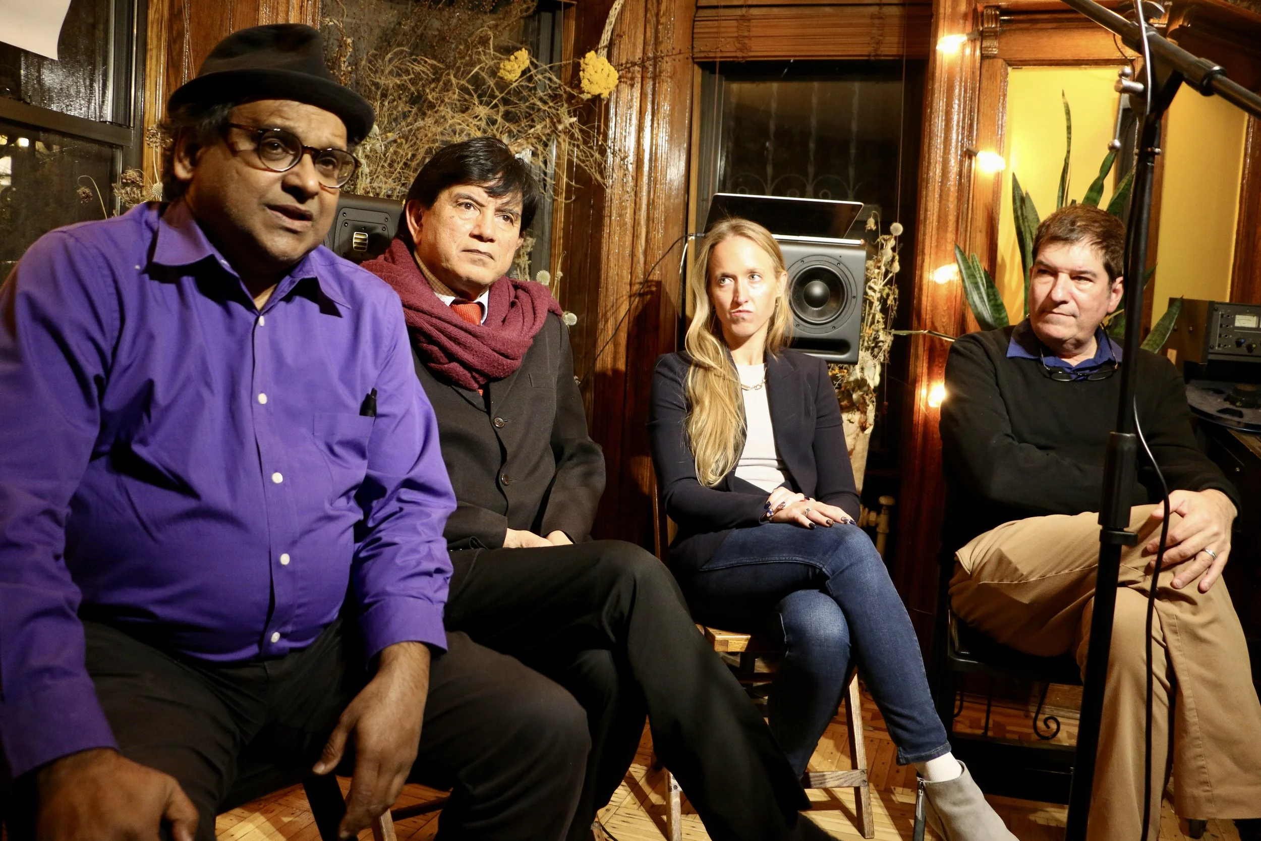 Group of four people sitting together in a cozy room, with wood paneling and speakers in the background. The person on the left wears a purple shirt and hat, the second person a black coat and maroon scarf, the third person a navy blazer, and the fou