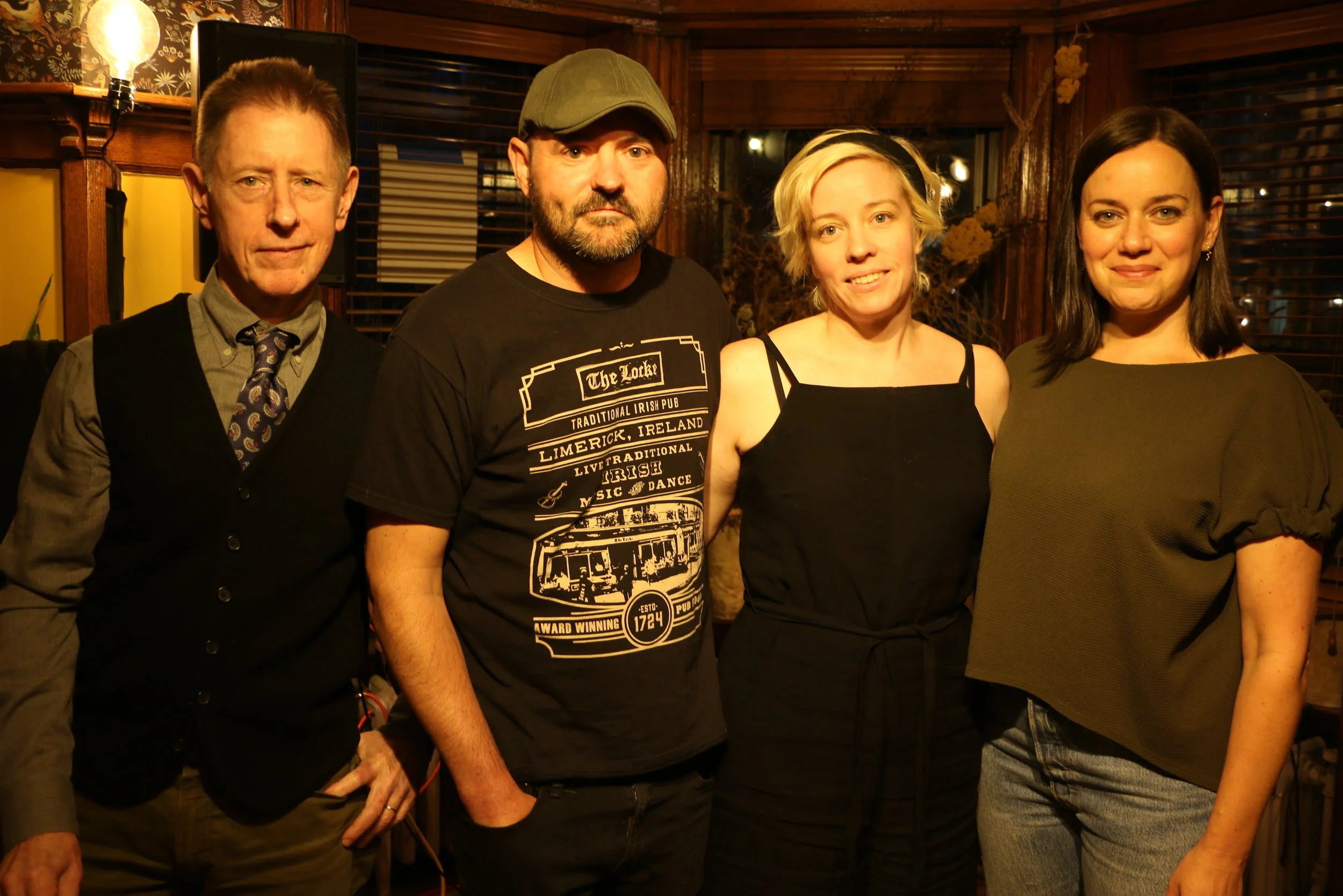 Four people posing together indoors, two men and two women. One man in a suit and vest, another in a black T-shirt and cap. One woman with short blonde hair, the other with long dark hair. Warm lighting and wooden interior decor.