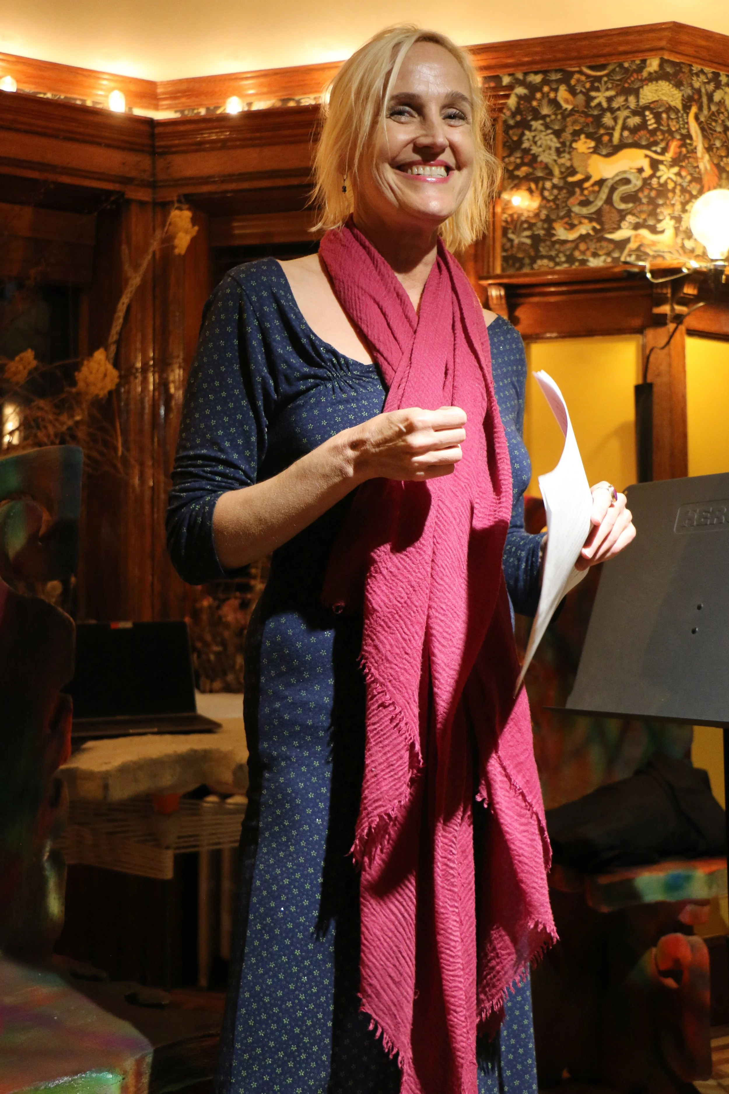 A woman smiling, wearing a blue dress and pink scarf, standing in a dimly lit, decorated room, holding a paper. The background features ornate wallpaper and wooden paneling.