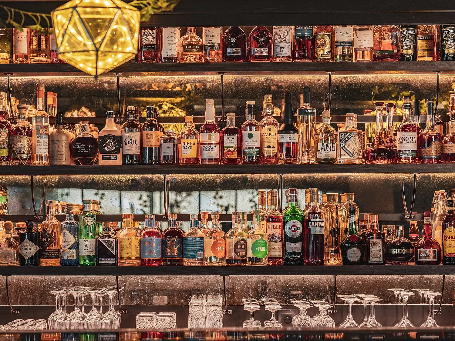 Étagère de bouteilles d'alcool variées dans un bar, avec verres en dessous et éclairage chaud.