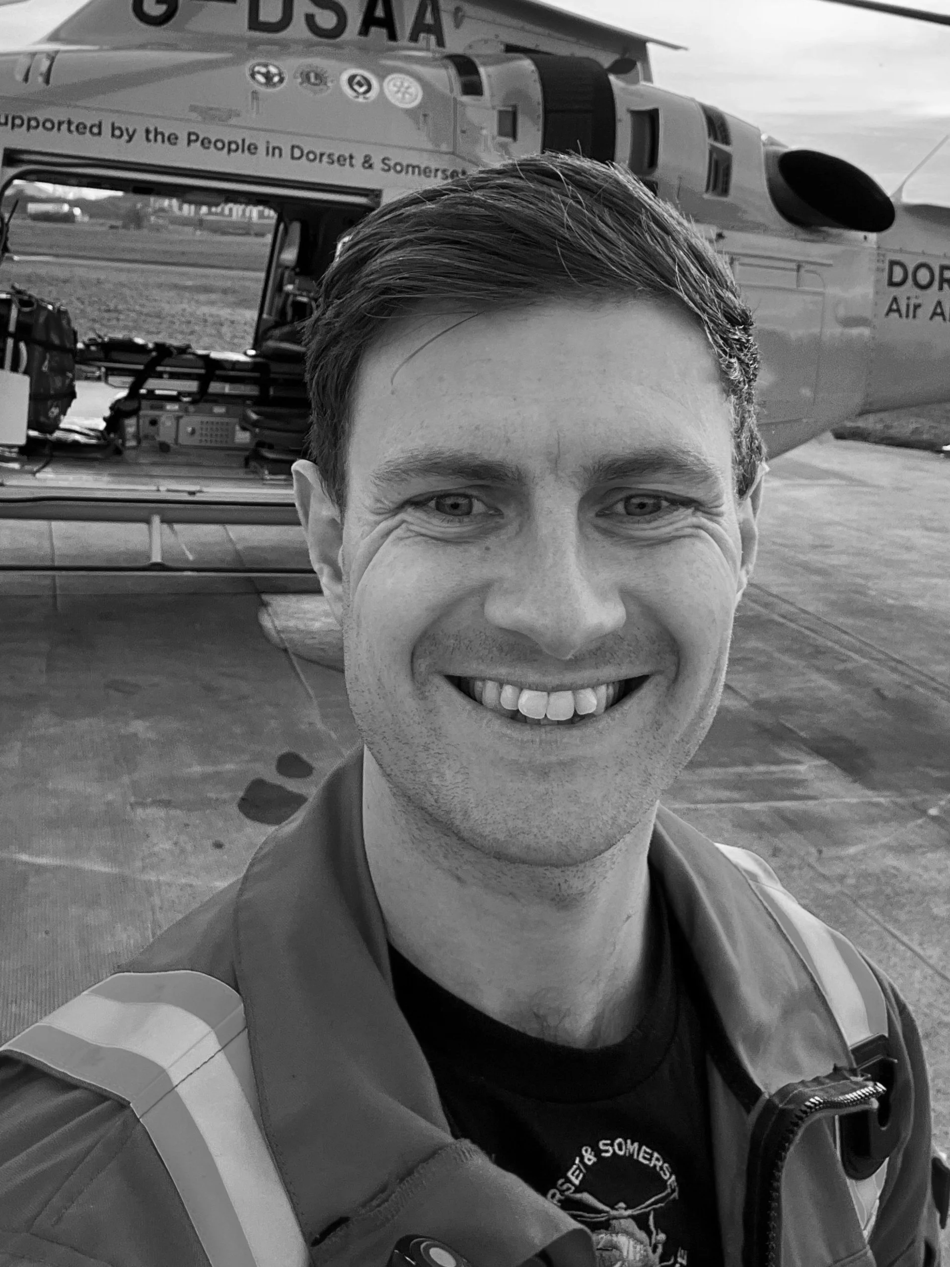 A man smiling in front of a helicopter labeled "Dorset & Somerset Air Ambulance." The helicopter appears open, with equipment visible inside. The man is wearing a jacket with reflective stripes.