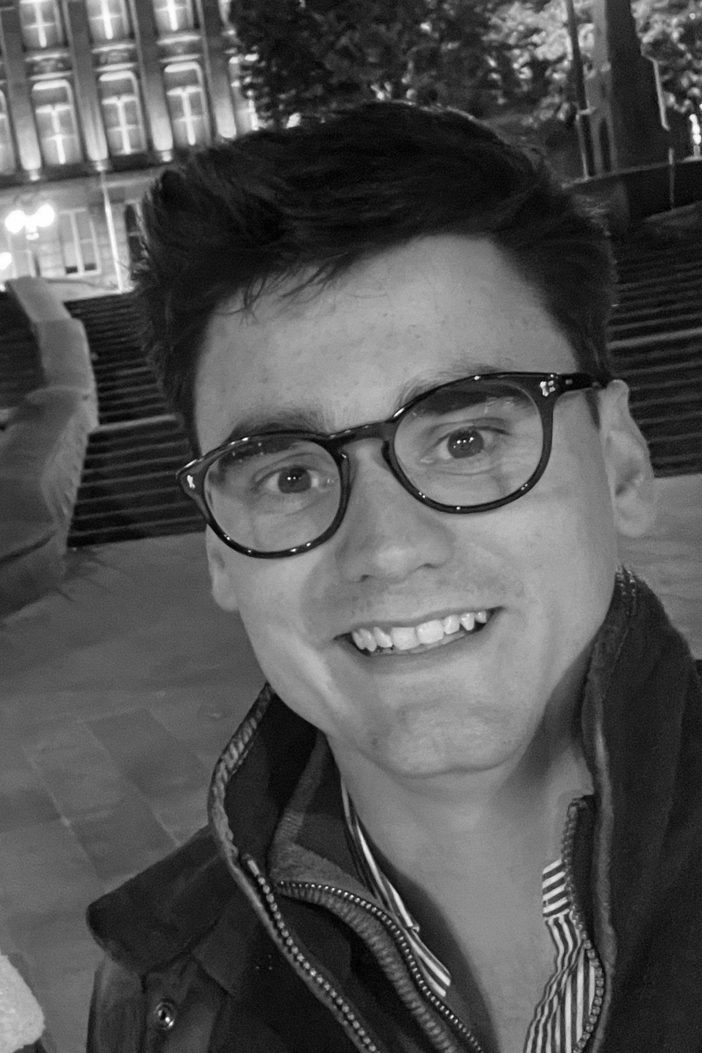 A smiling person wearing glasses and a jacket in an outdoor nighttime setting with buildings and steps in the background.
