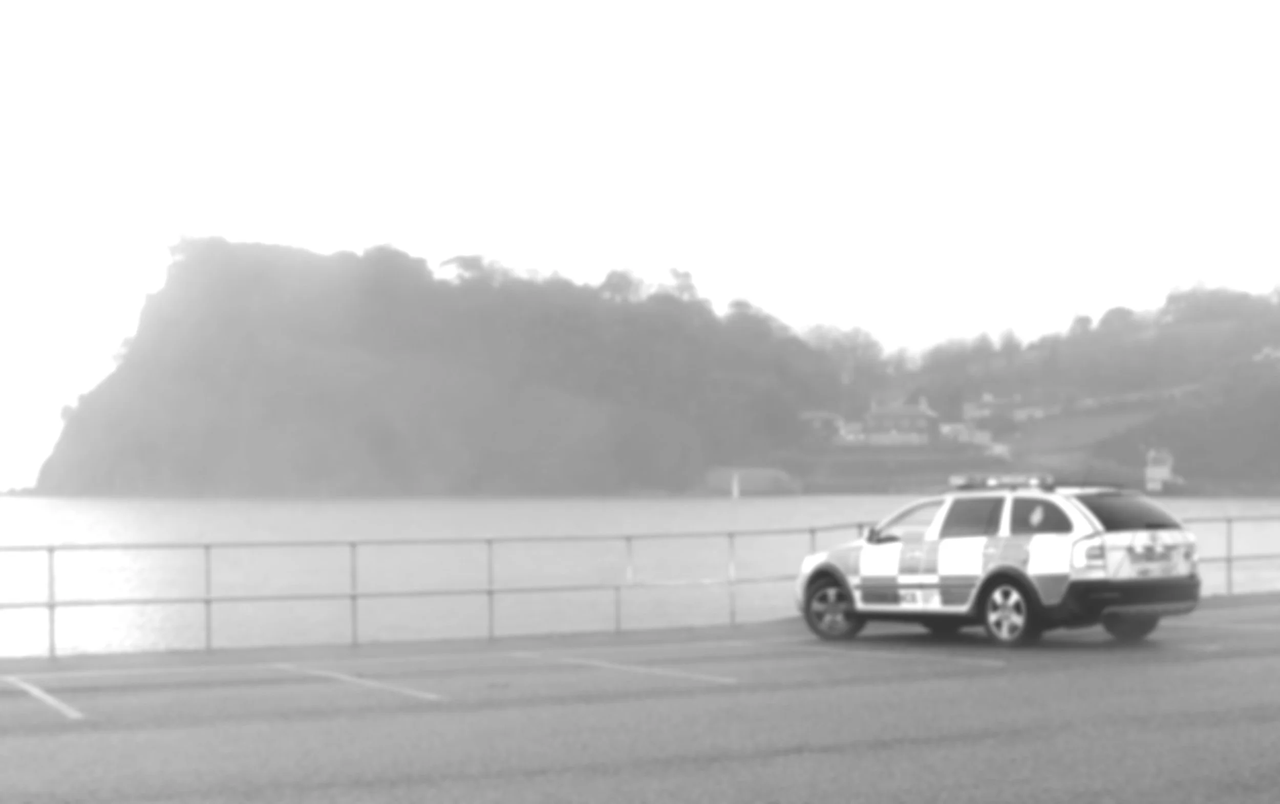 Police car parked near a waterfront with hills in the background.