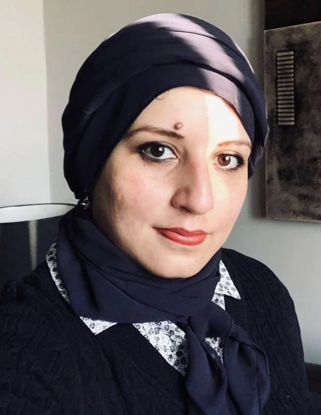 Portrait of a person wearing a dark headscarf and black sweater with a patterned collar, indoors with natural light. Ghazala