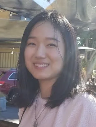 Person with dark hair smiling outdoors, blurred background