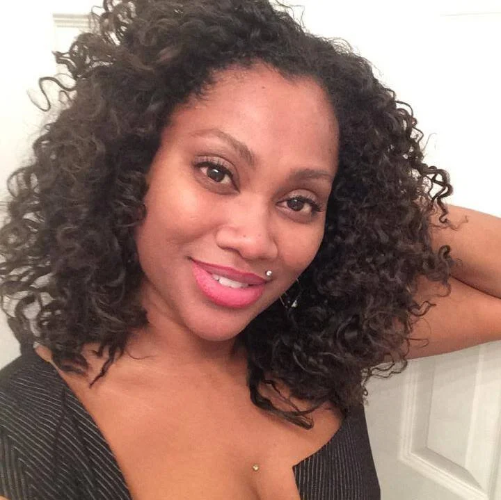 Smiling woman with curly hair and piercings, wearing a black top. Jeanelle