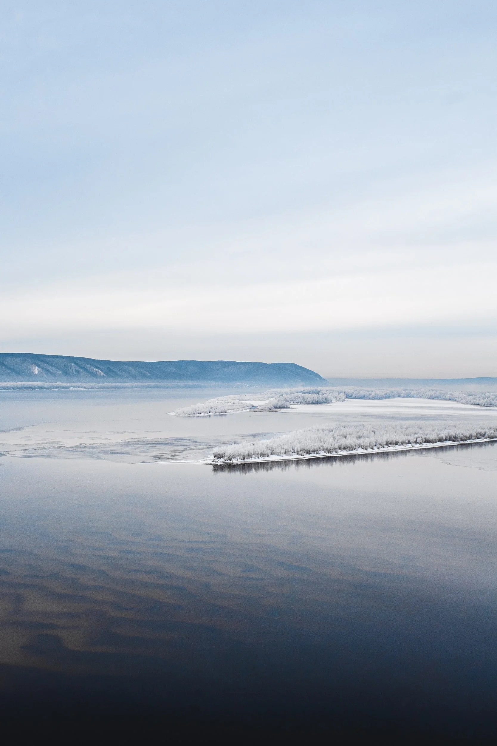 Snow-covered landscape with a calm body of water and distant snowy hills under a cloudy sky.
