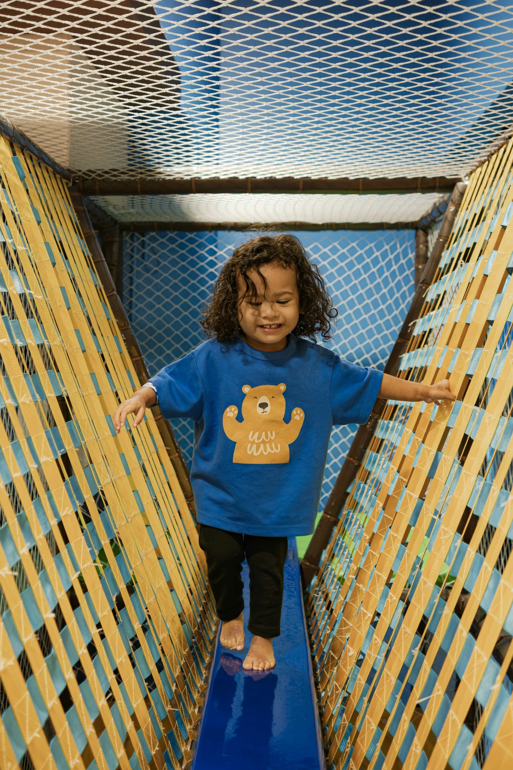 A young girl with curly hair wearing a blue shirt with a bear graphic walking on a small slide inside an indoor play area.