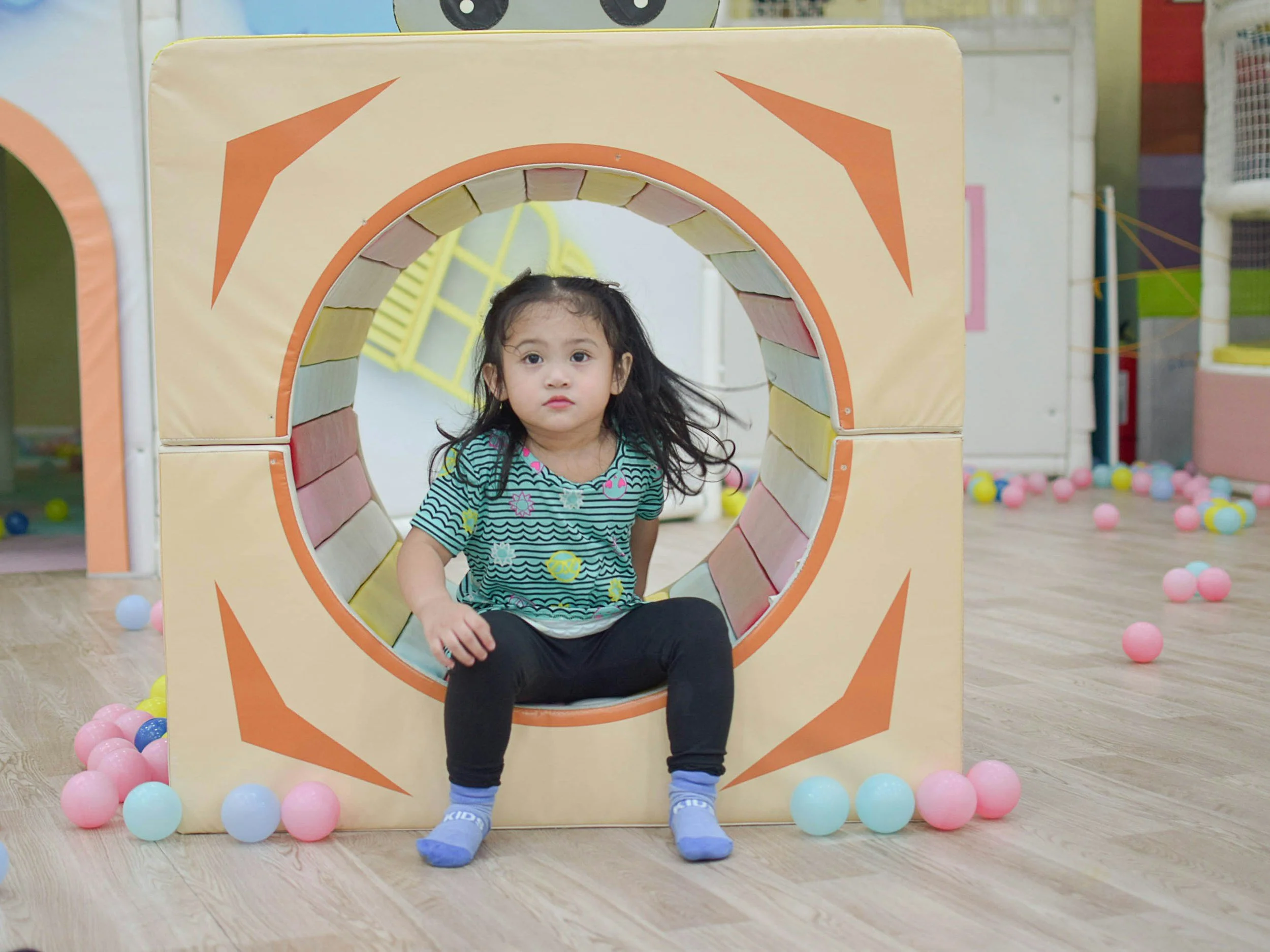 Young girl sitting inside a large, circular play tunnel with colorful balls scattered on the wooden floor around her, in an indoor play area.