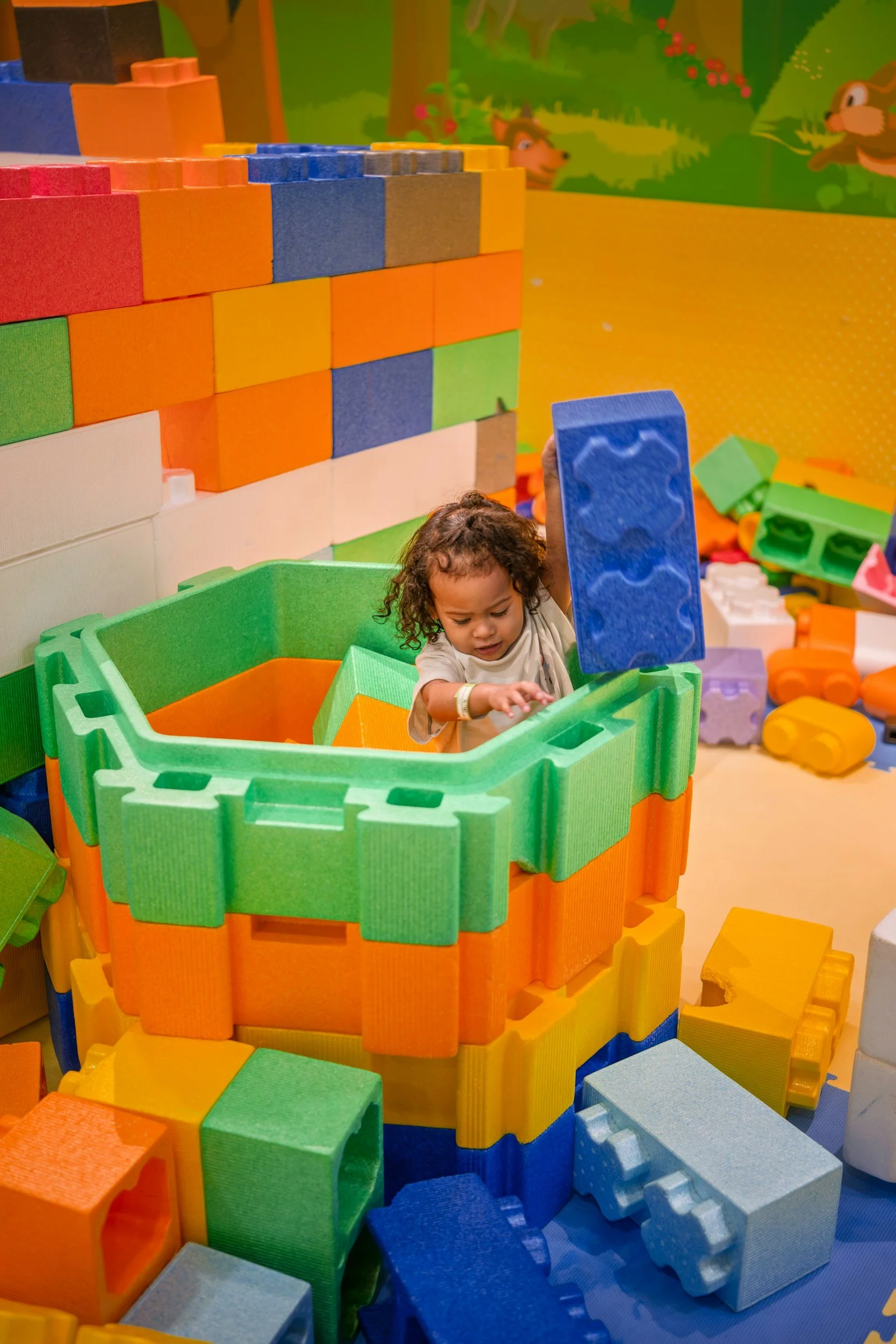 A young child, playing in a large indoor play area with colorful foam blocks, including a green and orange fortress-like structure, holding a blue foam block, with a mural of forest animals in the background.
