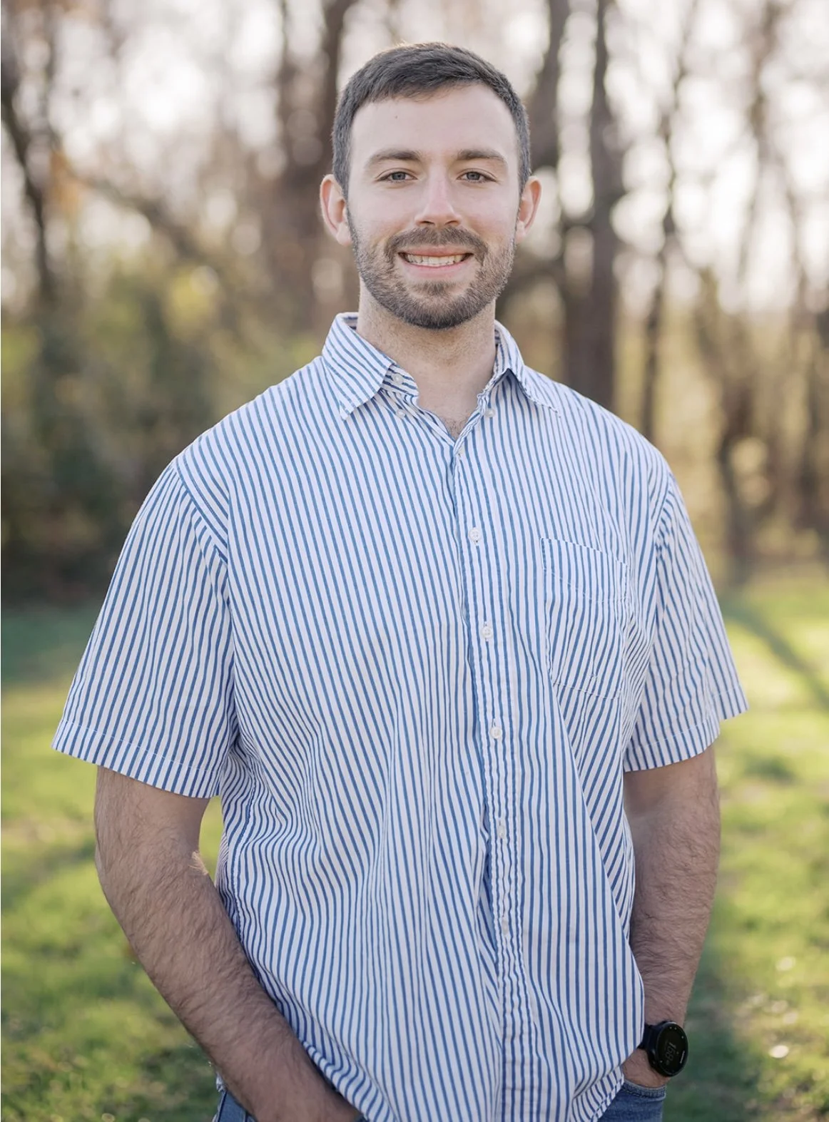 A young man with short dark hair, beard, and mustache smiling outdoors in a park during daytime, wearing a light blue and white striped button-up shirt, with trees and sunlight in the background.