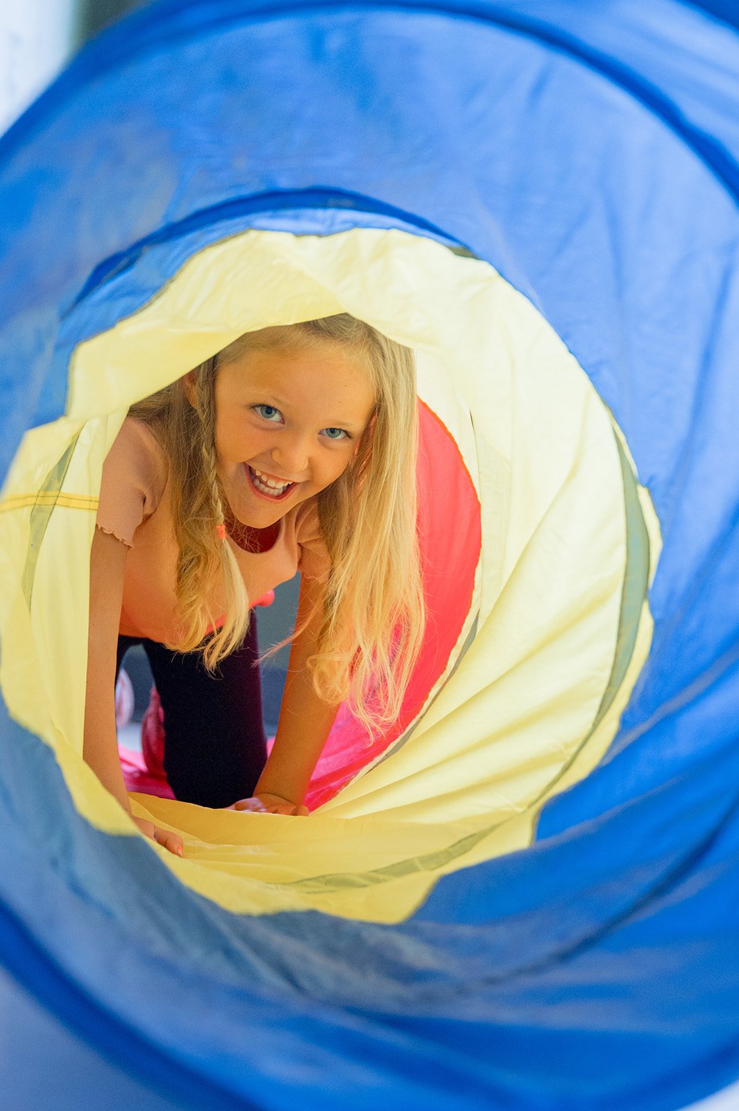 A young girl with curly hair wearing a blue shirt with a bear graphic walking on a small slide inside an indoor play area.
