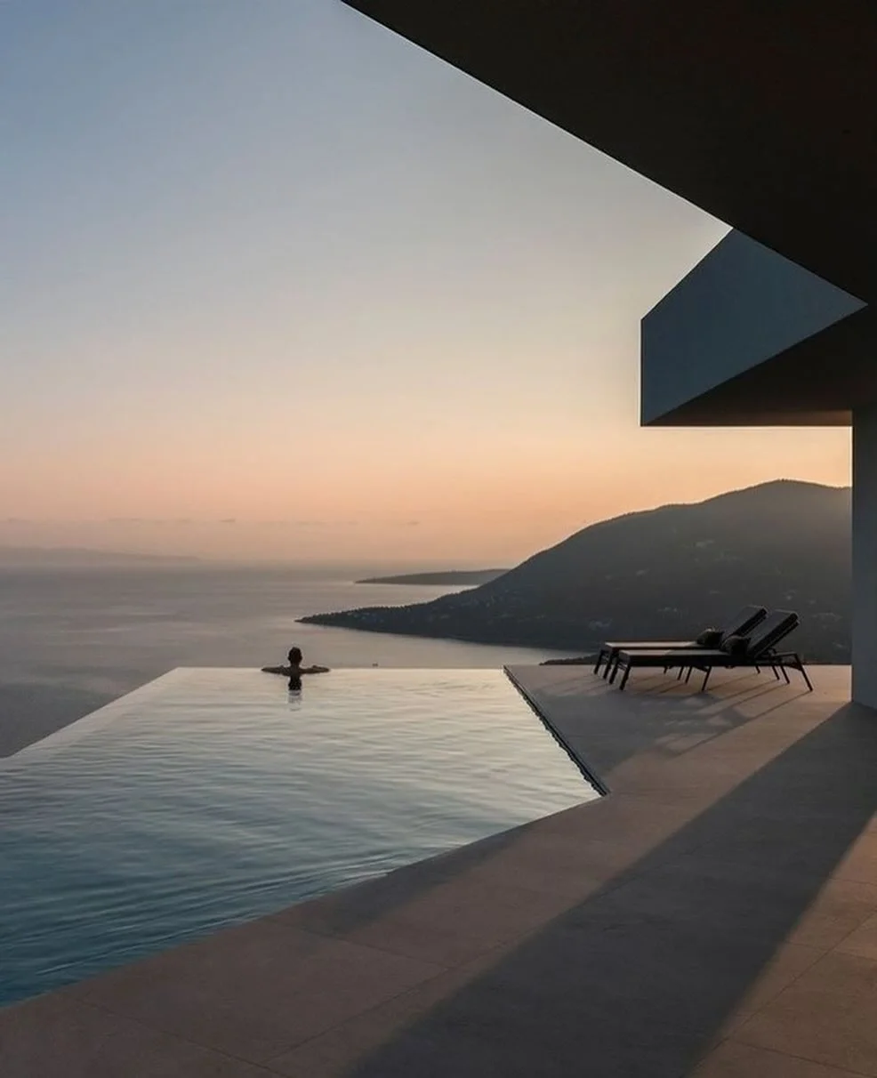 Persoon die in een infinity pool zit met uitzicht op de oceaan en bergen bij zonsondergang, naast twee ligstoelen op een terras.