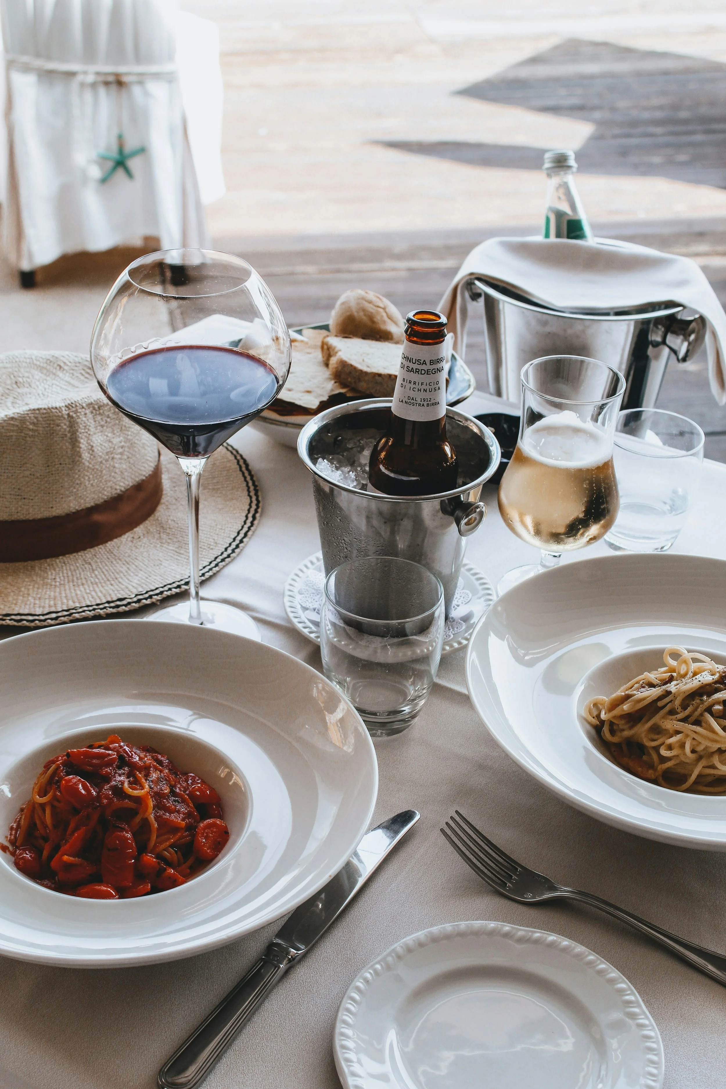 Voorbeeld van een feesttafel met pasta, wijn, bier en water. Midden op de tafel staan borden met pasta, een glas rode wijn, een glas witte wijn en een fles bier in een ijsemmer. Er ligt also een hoed en een bord met brood op de tafel.