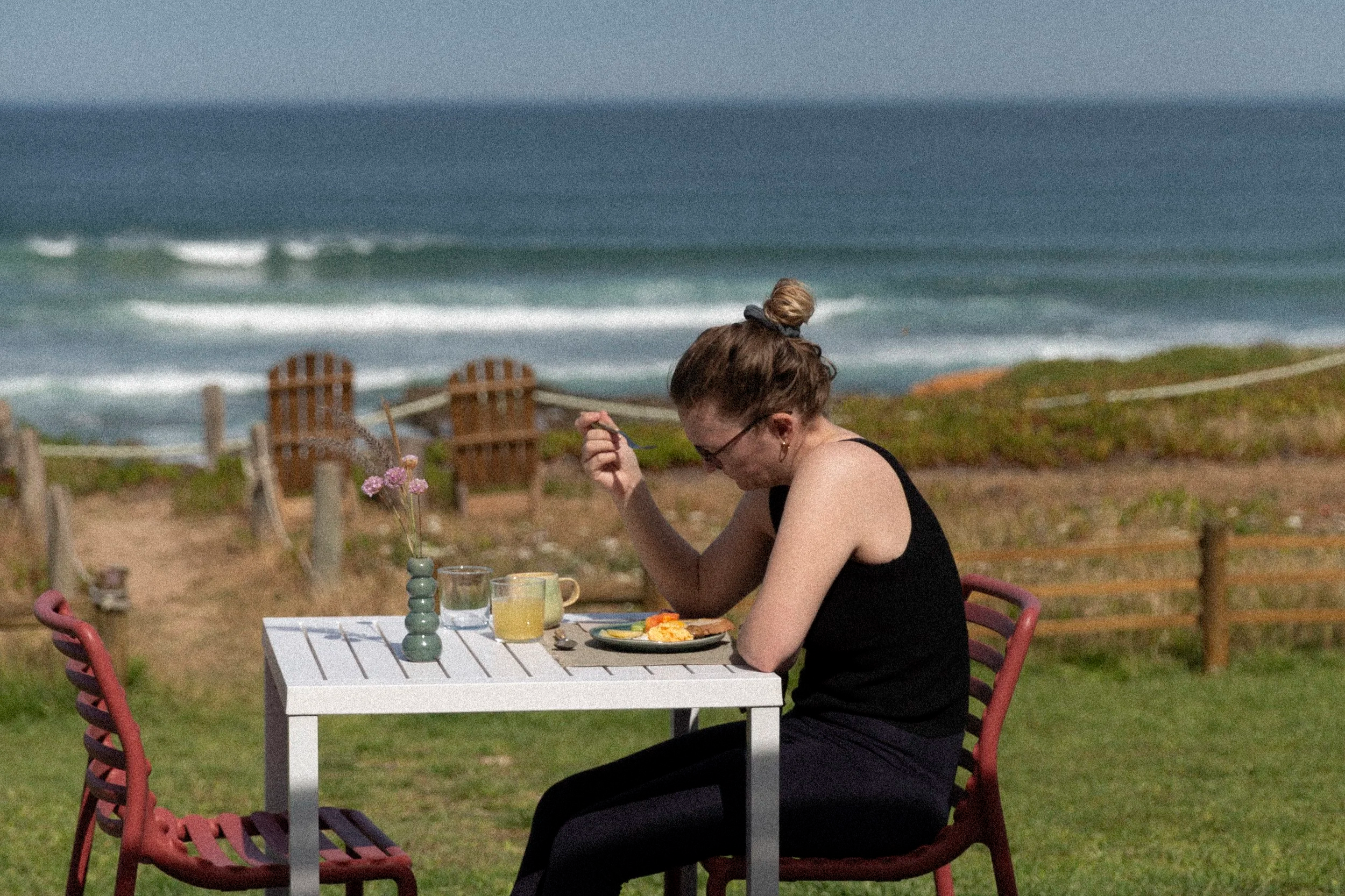 Eine Frau mit Brille und kurzen Haaren sitzt an einem weißen Gartentisch im Freien beim Essen, im Hintergrund ein Strand mit Wellen und Sitzgelegenheiten.