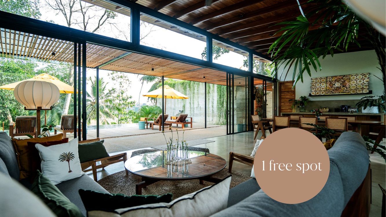Wohnzimmer und Terrasse mit offener Glaswand, moderne Einrichtung, Pflanzen, Holztisch, Sitzkissen mit Palmenmotiv, Sonnenschirm, Ausblick auf tropischen Garten.