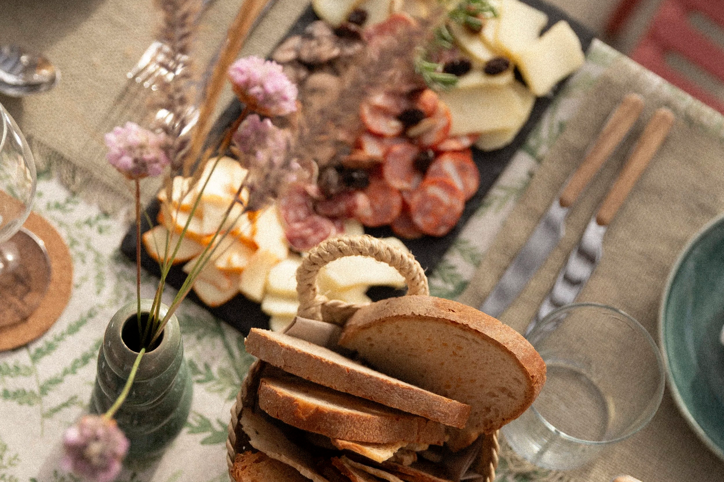 Eine Platte mit verschiedenen Käsesorten, Wurst, Oliven und Brot, serviert auf einem Tafelservice, mit einer kleinen Vase mit rosa Blumen