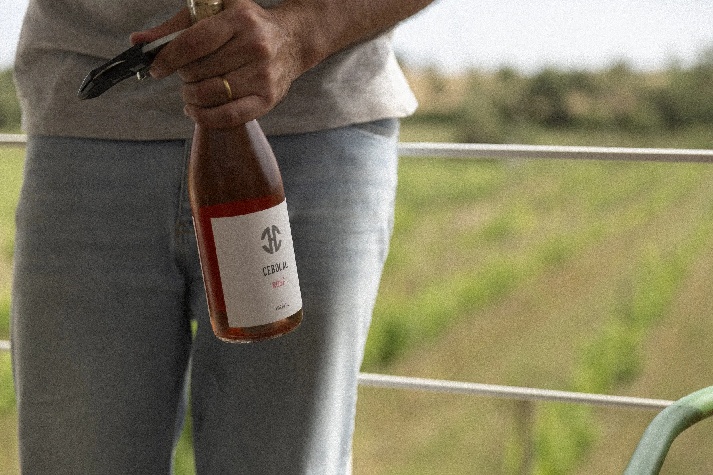 Nahaufnahme einer Person, die eine Flasche Rosé-Wein in der Hand hält, im Hintergrund ein grüner Garten oder Park. Die Person trägt eine goldene Ringe an der Hand und eine graue Hose.