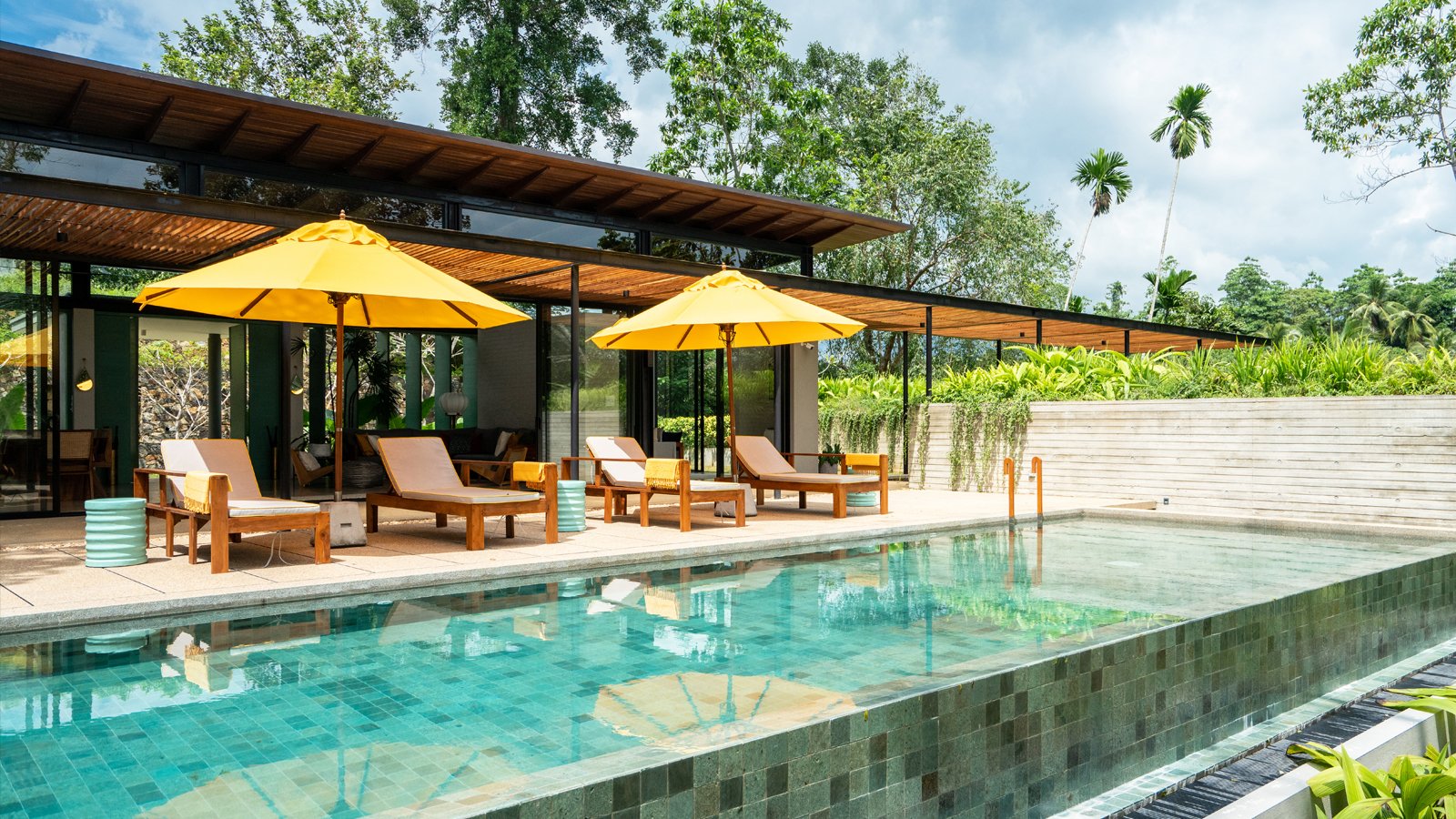Luxuriöse Villa mit Pool, Liegestühlen und gelben Sonnenschirmen, umgeben von tropischen Pflanzen.