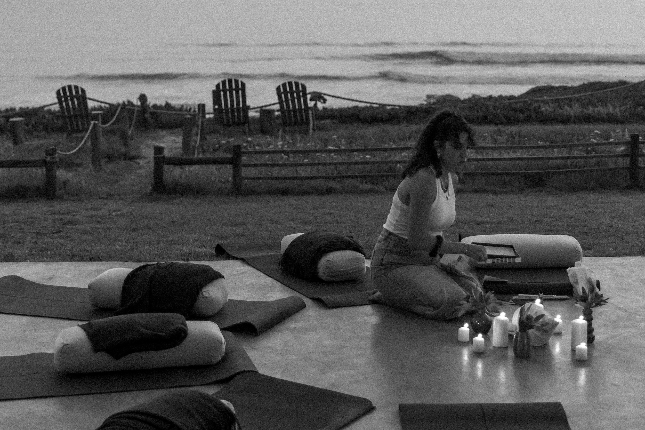 Andrea bei einer Yoga- oder Meditationssitzung im Freien im Abend- oder Nachtlicht, umgeben von Kerzen, mit einer Aussicht auf den Himmel mit Wolken im Hintergrund.
