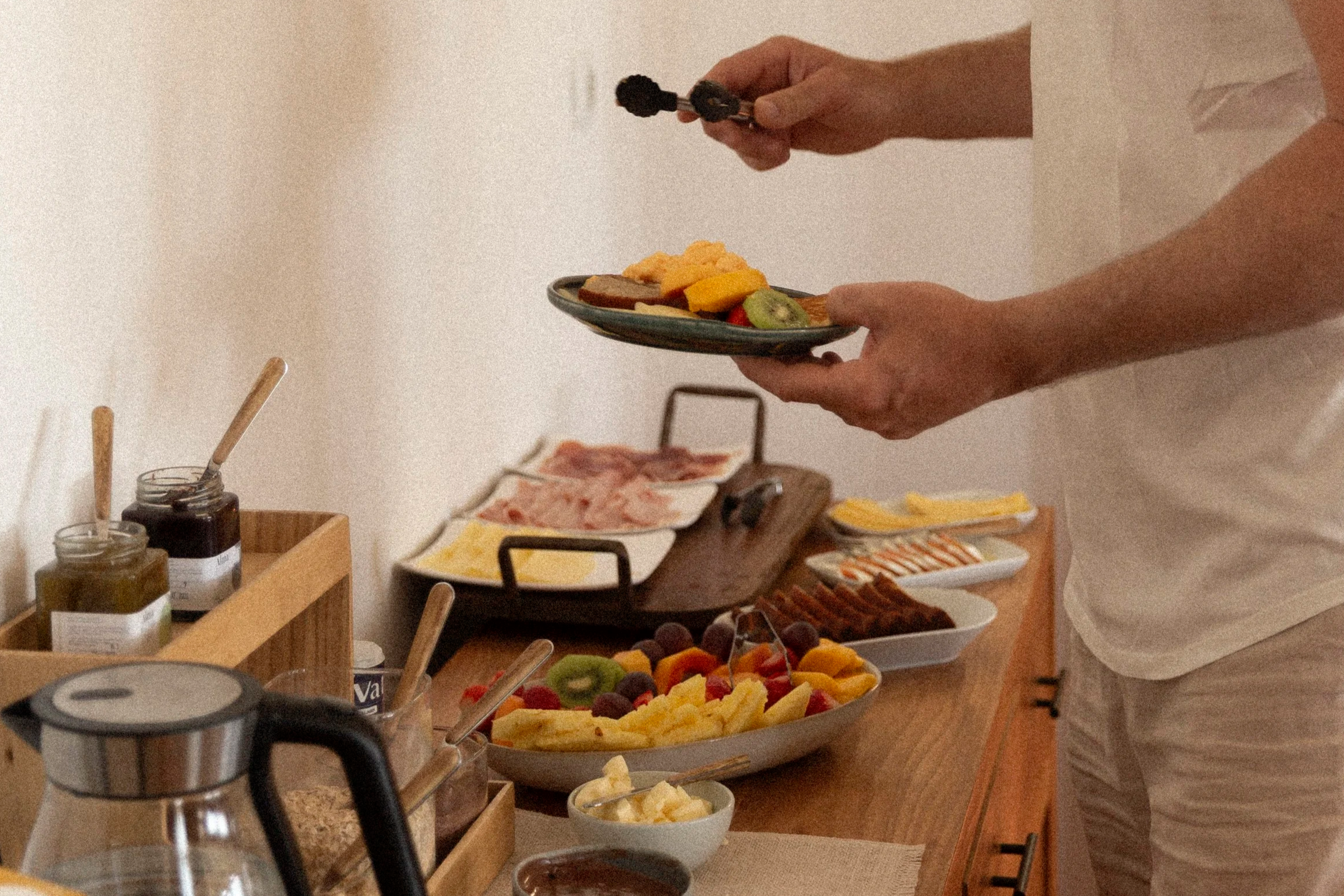 Person hält einen kleinen Teller mit Obst, darunter Kiwi, Ananas, Erdbeere und Brombeere, während im Hintergrund weitere Obst- und Wurstplatten auf einem Buffet liegen.