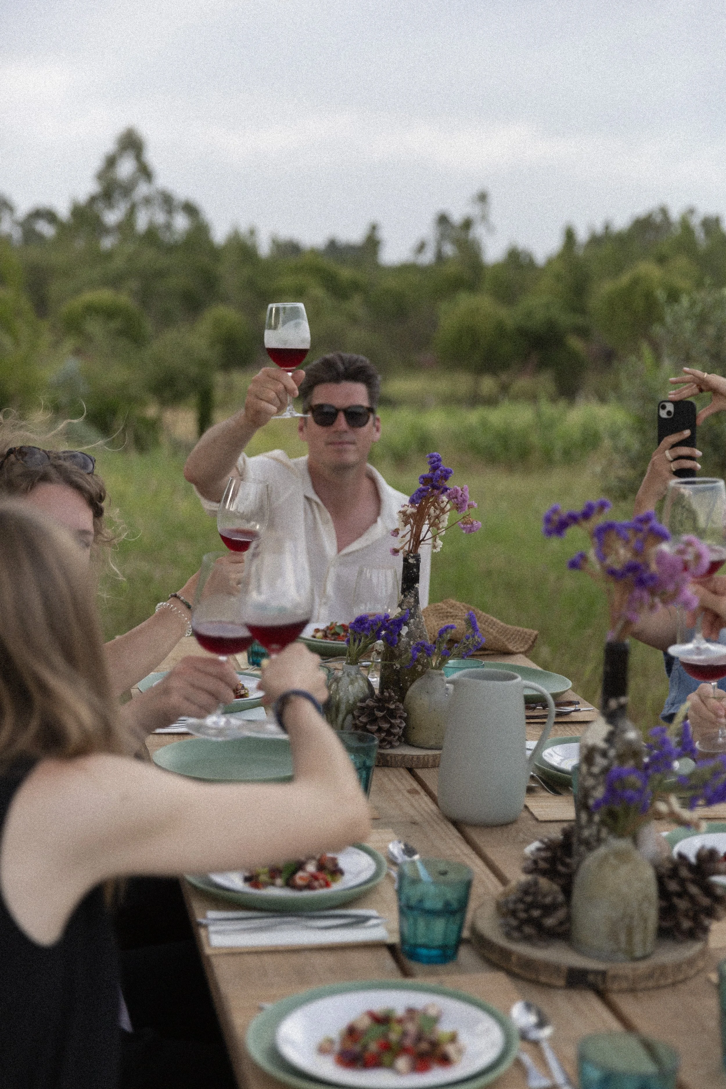 Menschen feiern ein Picknick im Freien mit Wein, Blumen und Essen auf einem Holztisch, umgeben von Natur und Bäumen im Hintergrund.