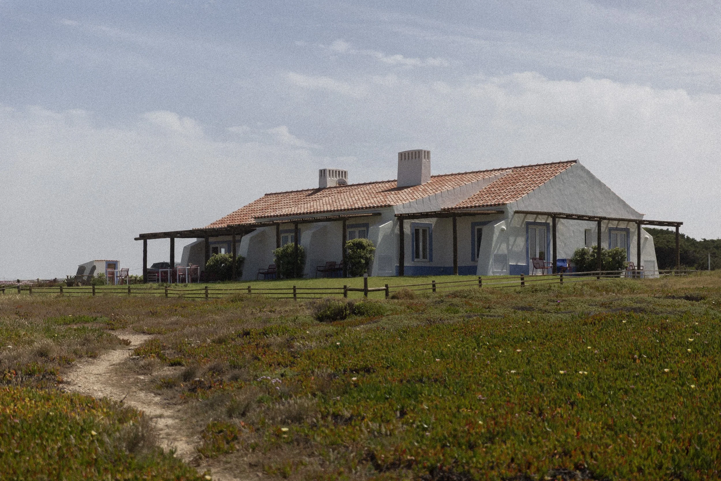 Ein Haus mit roten Ziegel-Dächern und weißen Wänden steht auf einer Wiese vor einem blauen Himmel mit wenigen Wolken.