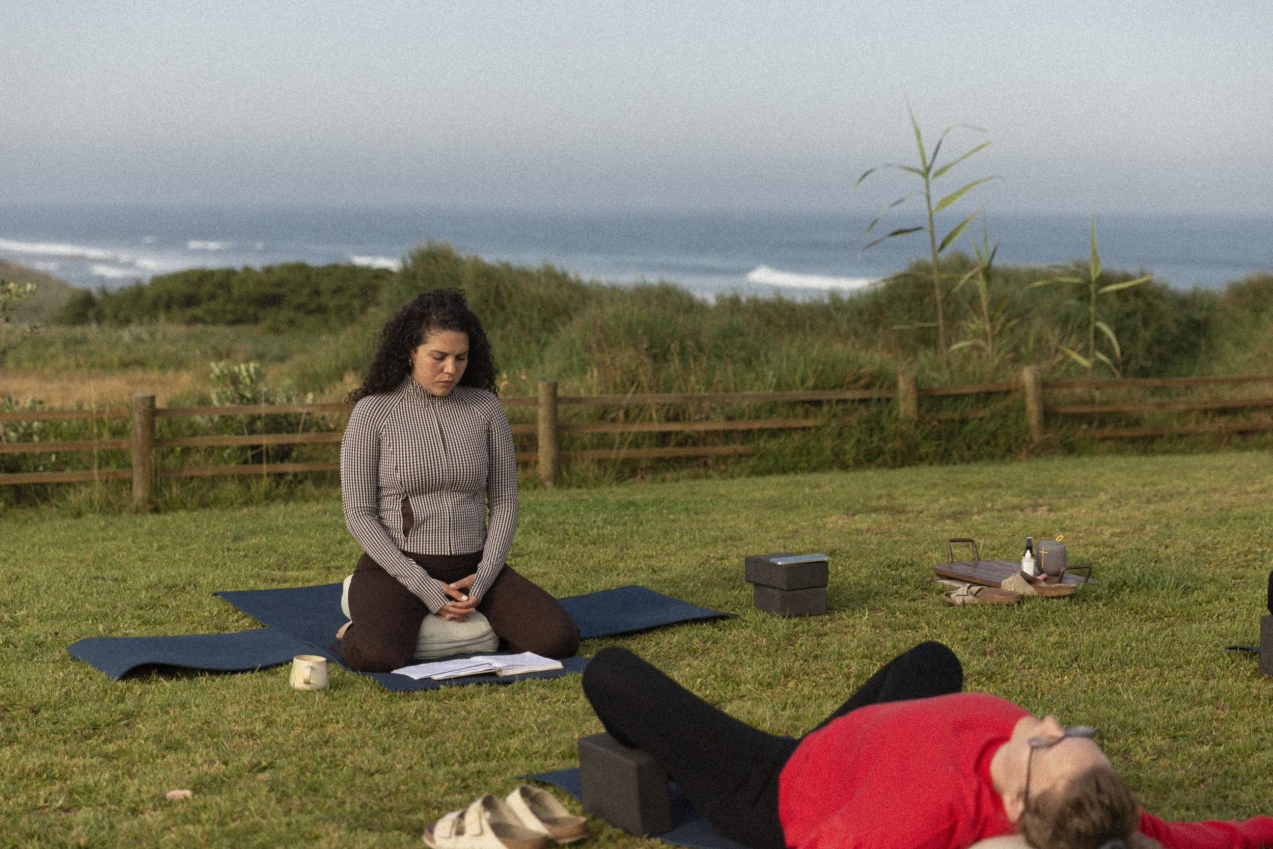 Eine Frau sitzt im Schneidersitz auf einer Matte im Freien, während im Vordergrund ein Mann auf dem Bauch liegend entspannt. Es sieht nach einer Yoga- oder Meditationssitzung aus, mit einem Rucksack, Tee und anderen Gegenständen auf dem Boden.