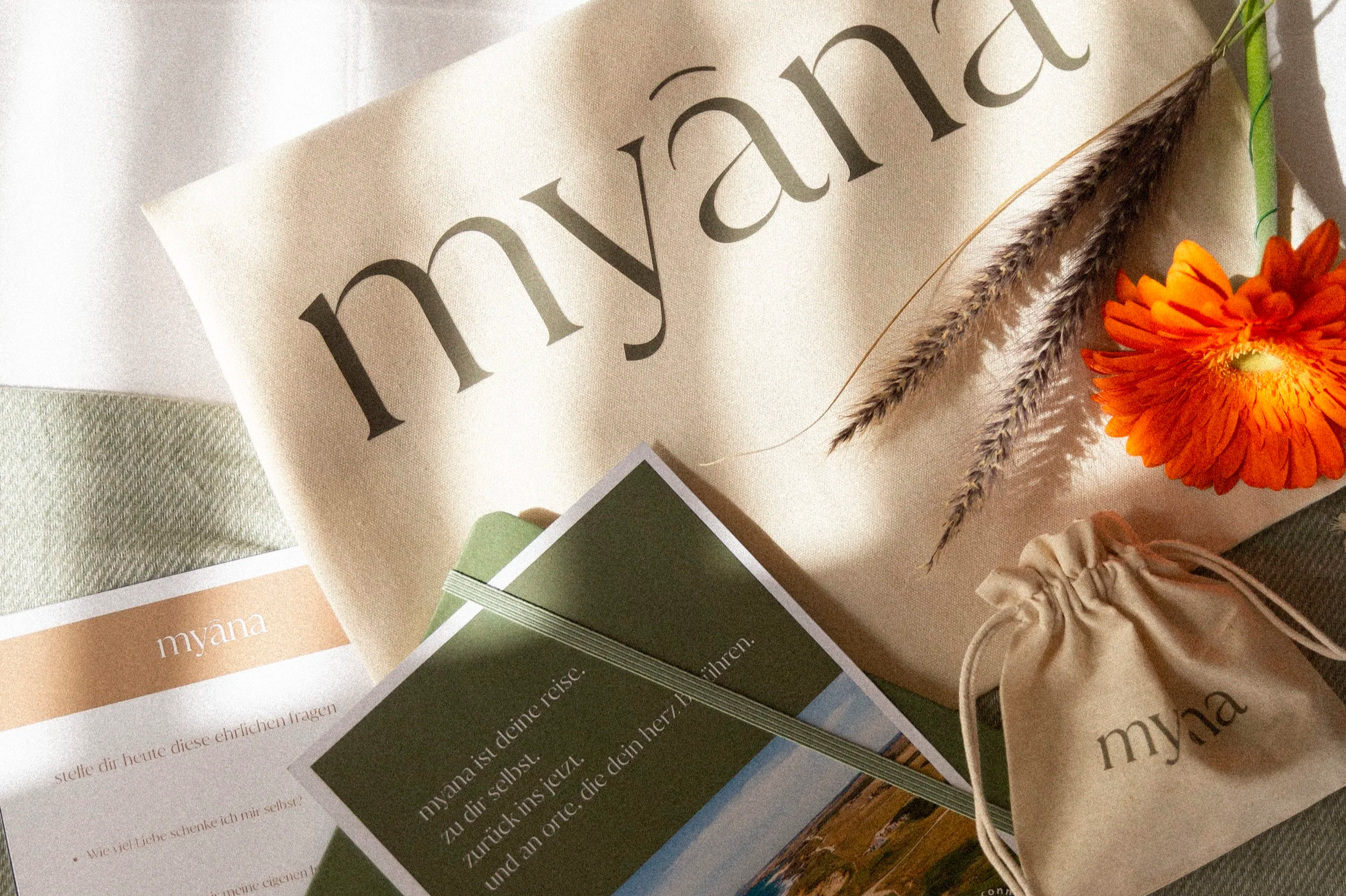 Blätter, eine orange Blume, Karten mit Text, ein Stoff mit dem Wort 'myana' in Grau auf Beigefarbenen Hintergrund.