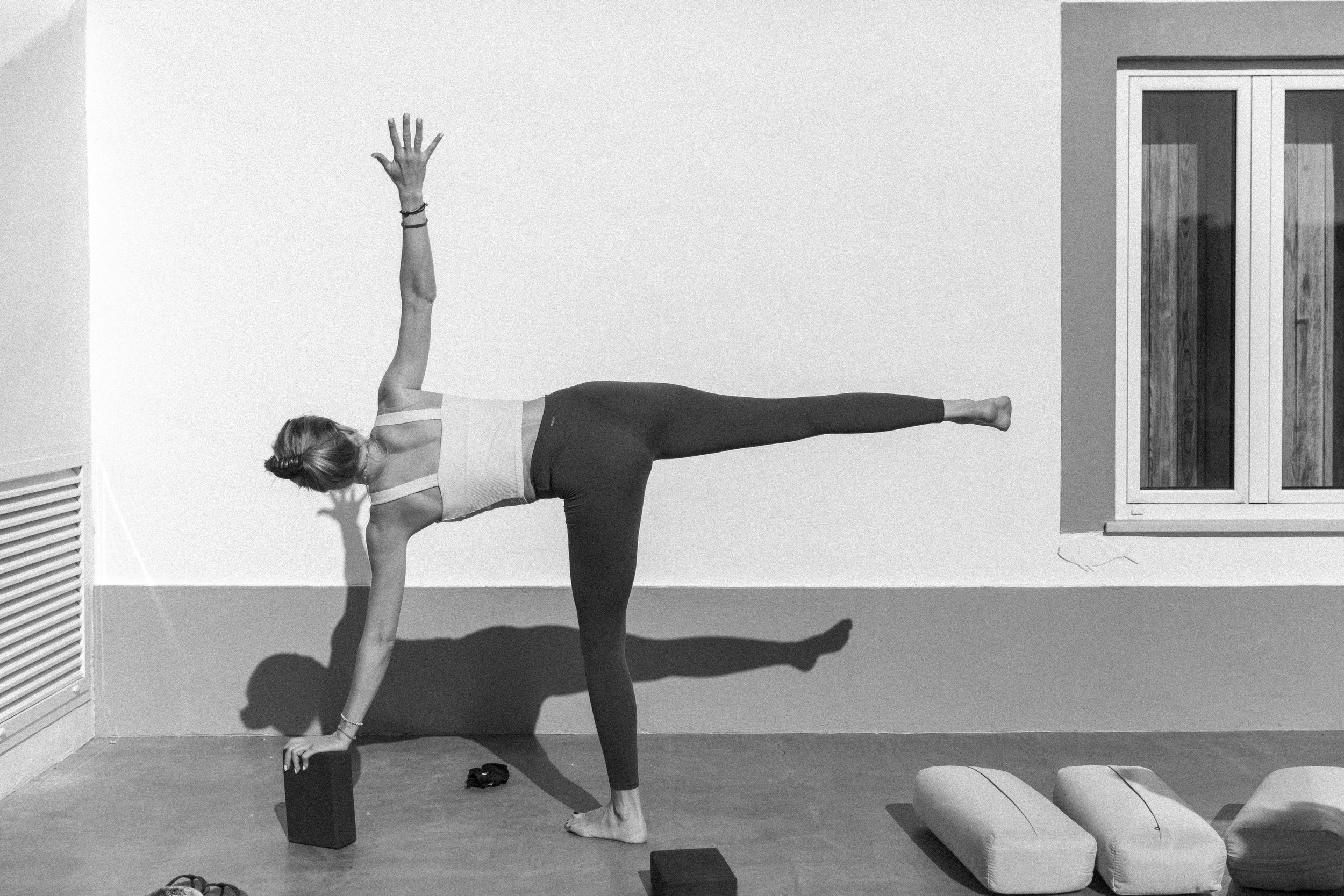 Frau macht Yoga in einem Raum mit weißen Wänden und einer Heizung auf der linken Seite, sie befindet sich in der Seitwärtsposition auf einem Yoga-Block, trägt eine weiße Sport-BH, schwarze Leggings und hat einen Haarfilm.