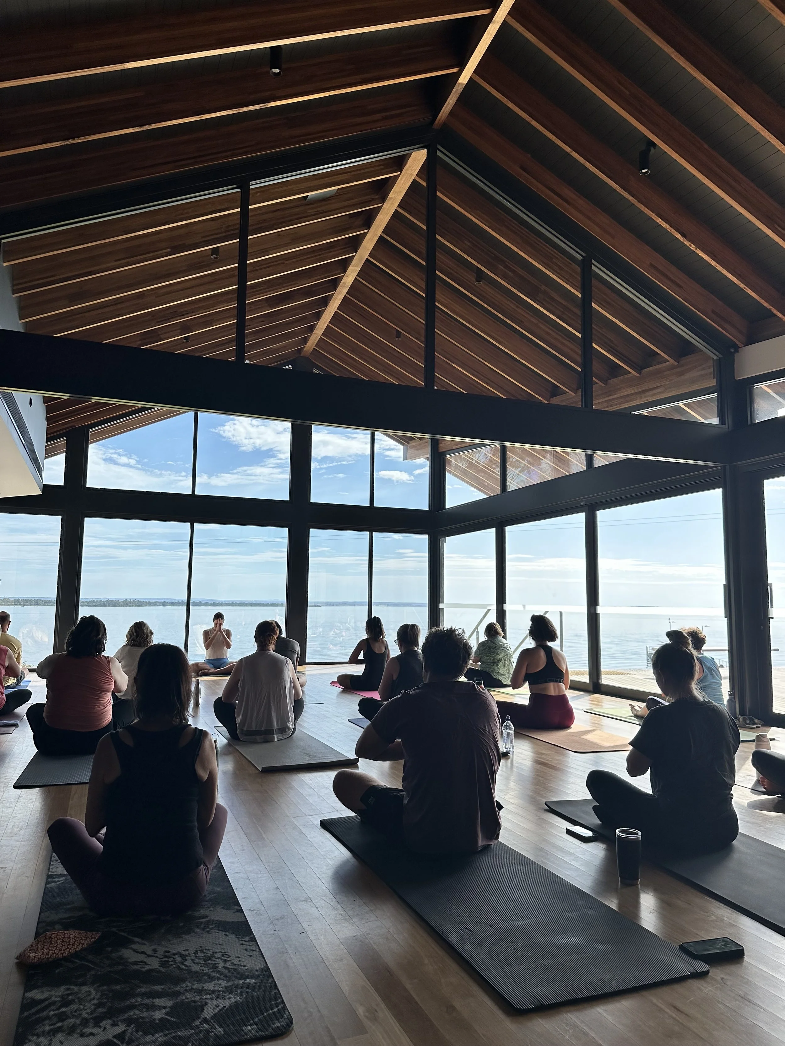 People participating in a yoga class in a spacious, sunlit room with large windows overlooking a body of water. Eagle Point, East Gippsland, near Paynesville and Bairnsdale.