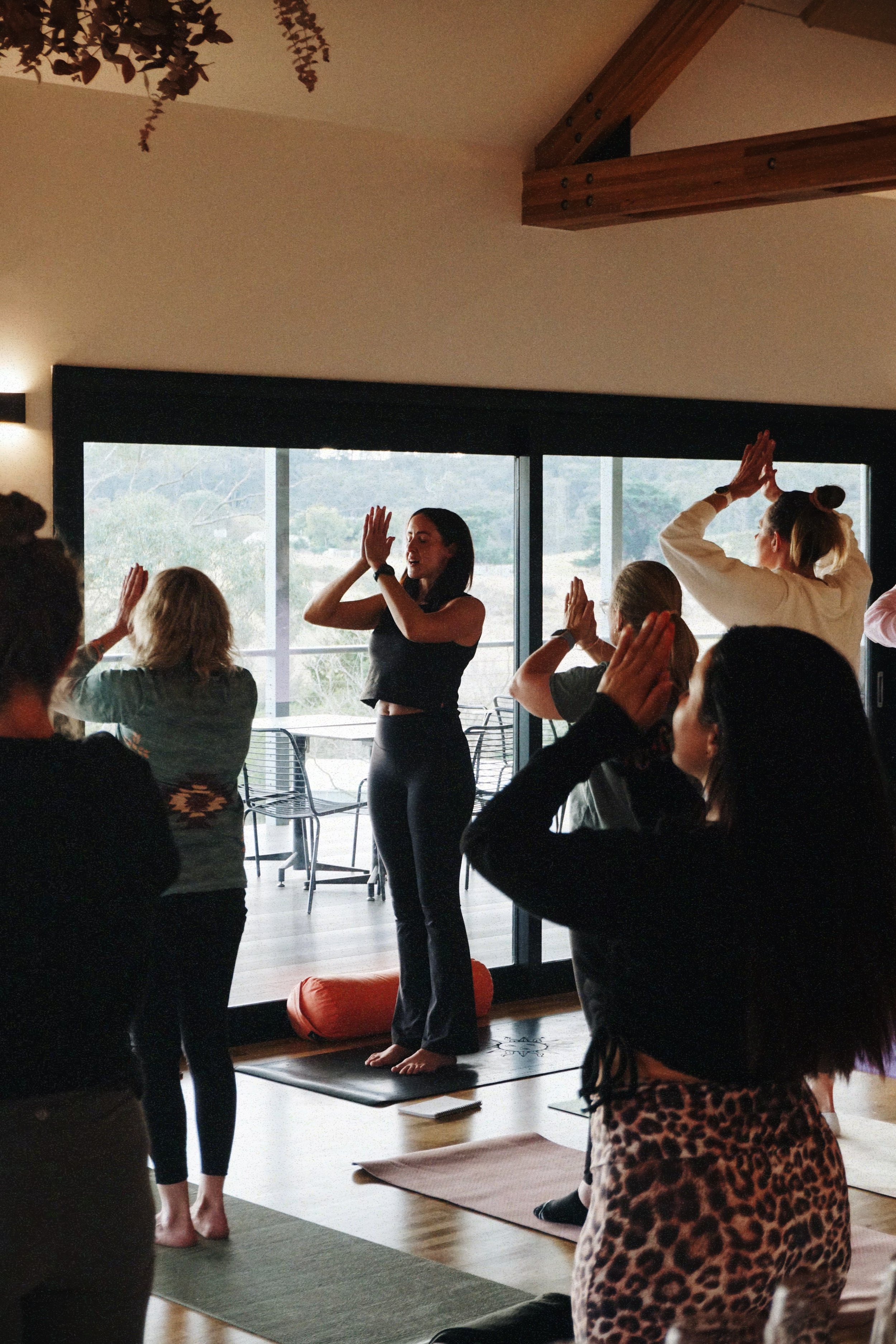 Group of women participating in a yoga class indoors, with a woman leading the session near a large window showing an outdoor view. A yoga event in Nicholson, East Gippsland, Victoria.