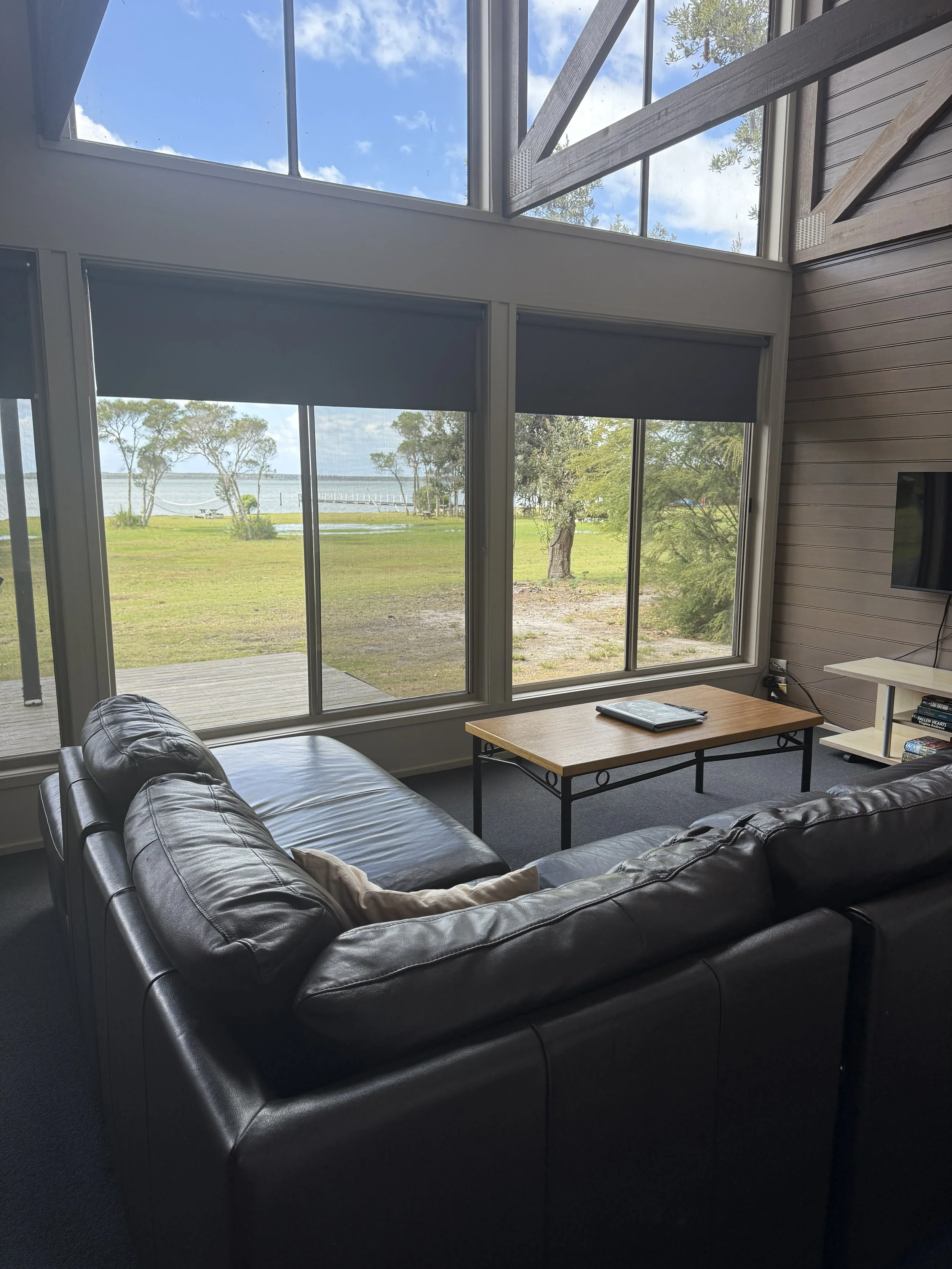 Living room with large windows overlooking a grassy yard and a body of water, brown leather sofa, wooden coffee table with a tablet, and a white media stand with a TV and books. A yoga retreat venue, Waterfront retreat Wattle Point in East Gippsland.