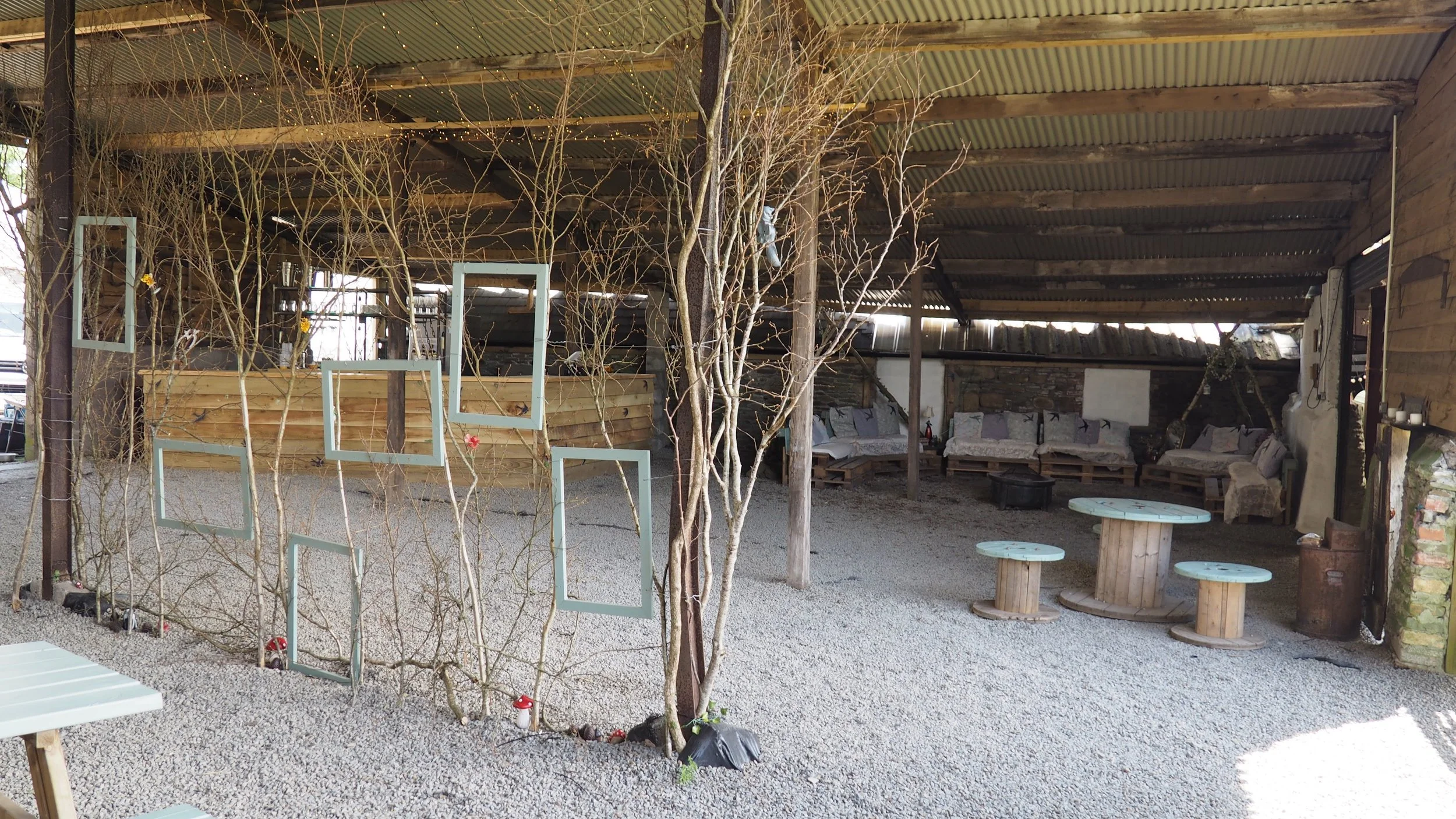 Indoor rustic seating area with gravel floor, wooden furniture, and leafless trees decorated with small flowers and framed mirrors.