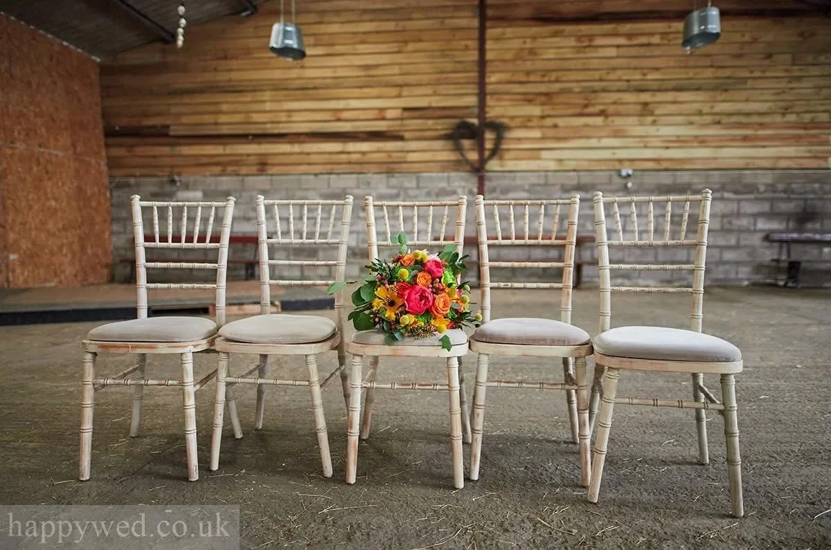 Five wooden chairs with beige cushions arranged in a row inside a rustic indoor space, with a colorful flower bouquet placed on the middle chair.