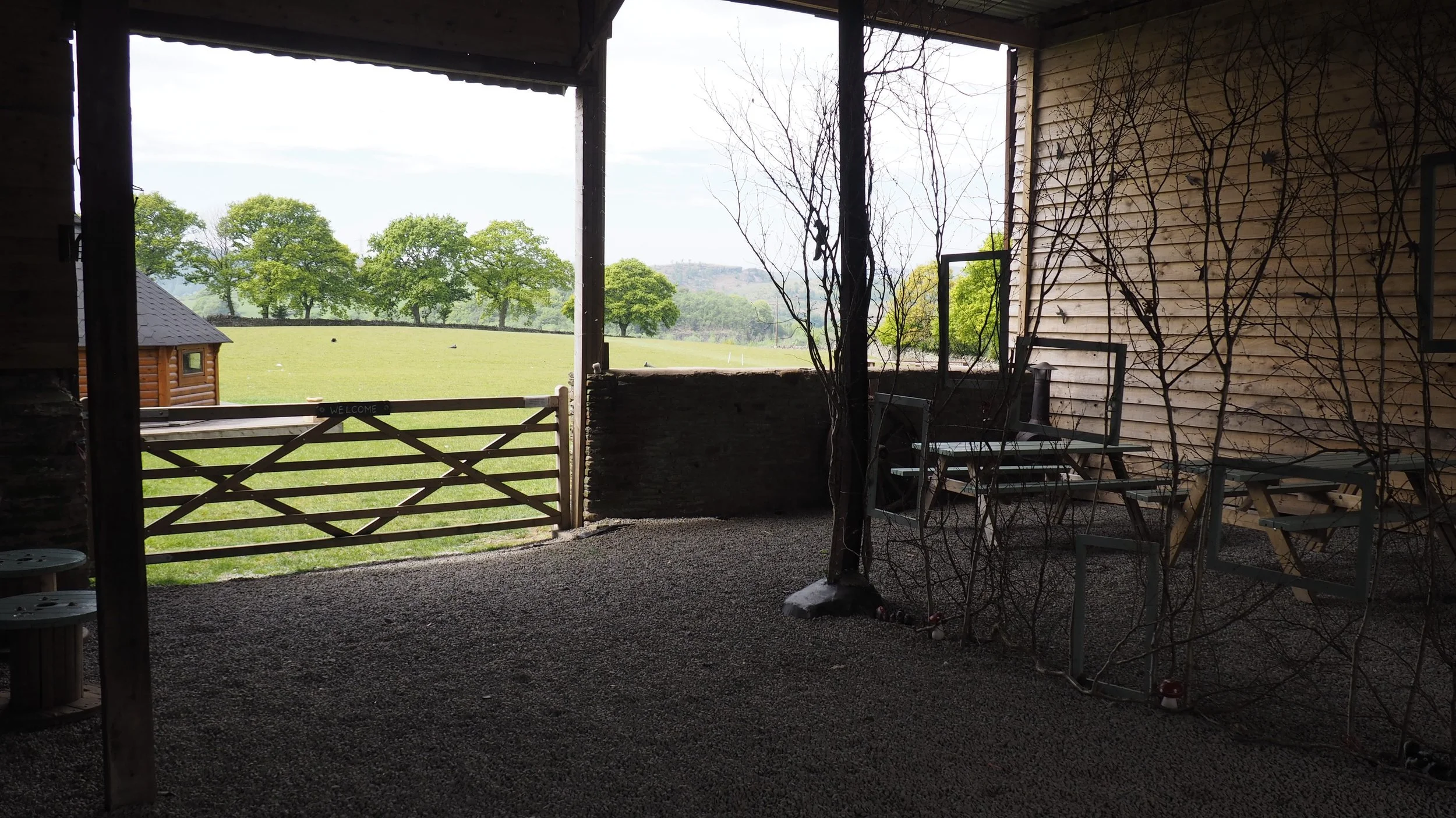 View from inside a rustic porch looking out over a grassy field with trees, a small log cabin, and distant hills.