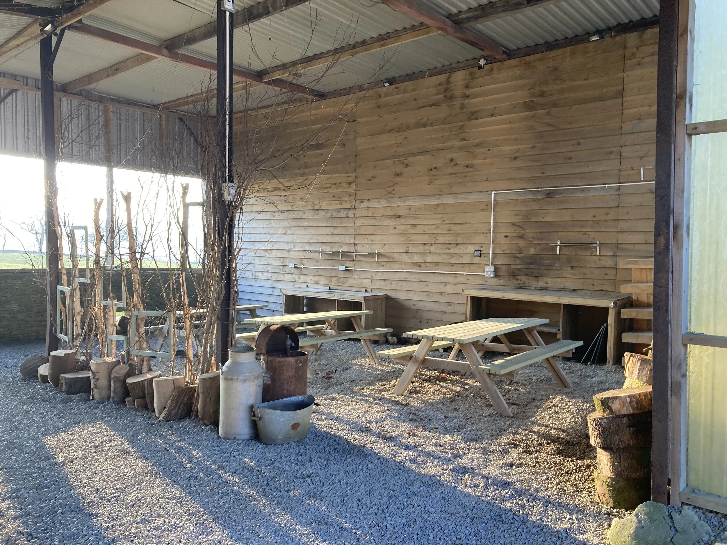 Outdoor seating area with wooden picnic tables under a metal roof, surrounded by tree branches and various containers, with gravel ground and a wooded area in the background.