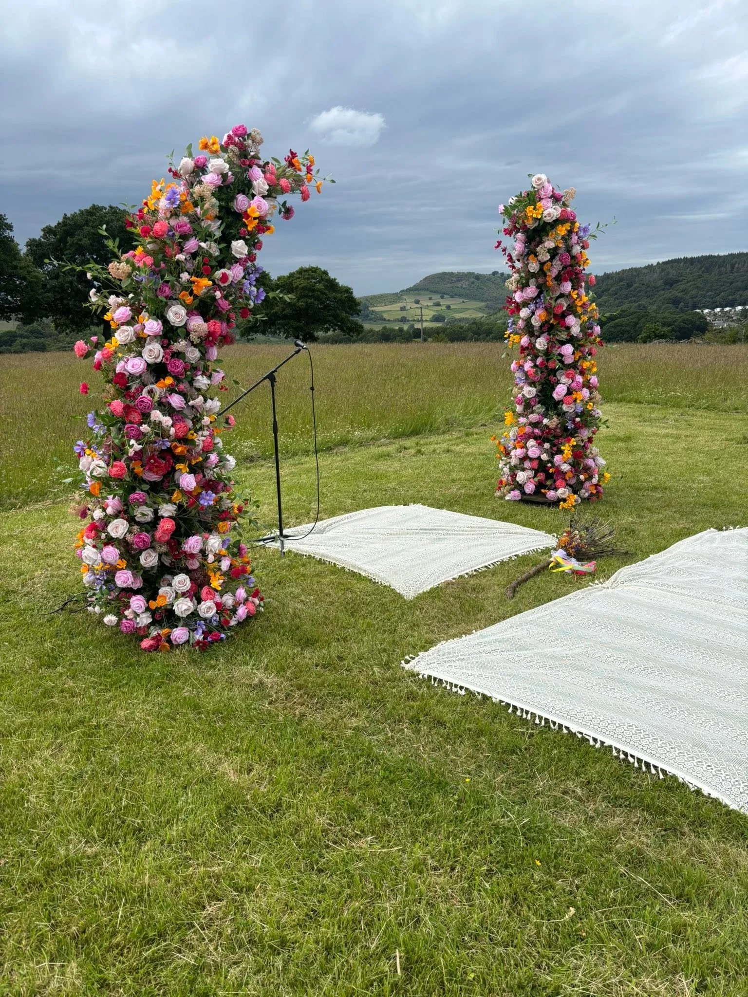 Two tall flower arrangements made of pink, purple, orange, and white flowers stand outdoors on a grassy field with rolling hills and a cloudy sky in the background, and microphone stands in front of them.