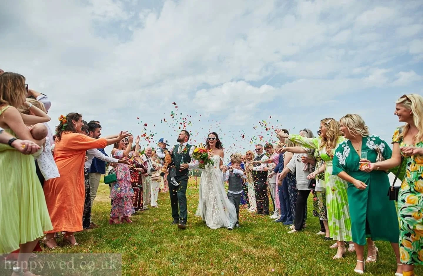 Wedding celebration outside with a bride and groom walking hand in hand amid guests throwing confetti, on a grassy field under a cloudy sky.