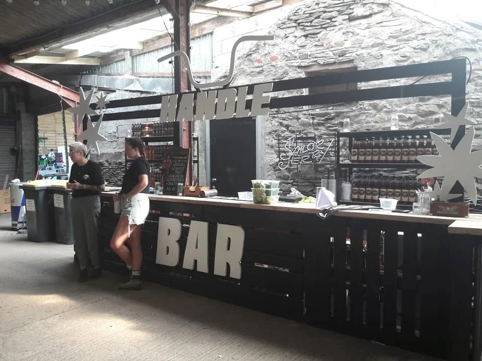 Indoor bar setup with a large black sign that reads 'HANDLE BAR' and additional text 'SALOIR SERVICES.' Two staff members stand behind the bar, with supplies and bottles displayed on shelves behind them. The setting appears lively and industrial.