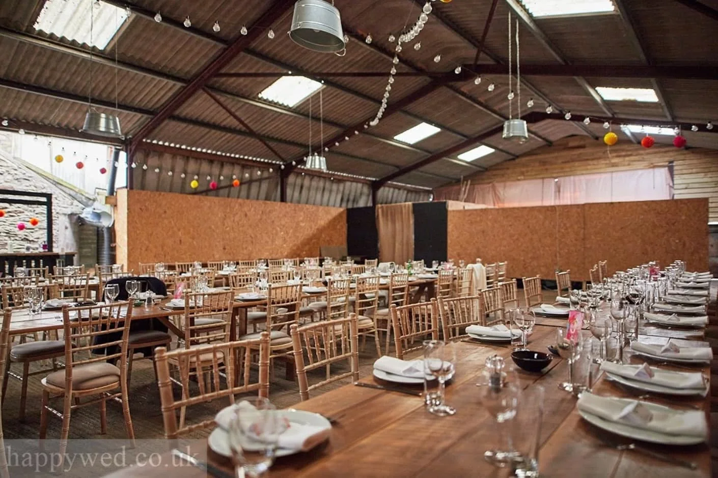 Large rustic event space decorated for a celebration with long wooden tables set with plates, glasses, and napkins, surrounded by wooden chairs, with string lights and colorful hanging decorations on the ceiling.