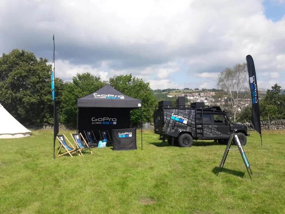 A GoPro promotional setup on a grassy field with a black canopy tent, promotional flags, foldable chairs, and a black van. The setup includes branding with the GoPro logo on the tent, flags, and chairs, and there is a scenic background with trees and a cloudy sky.