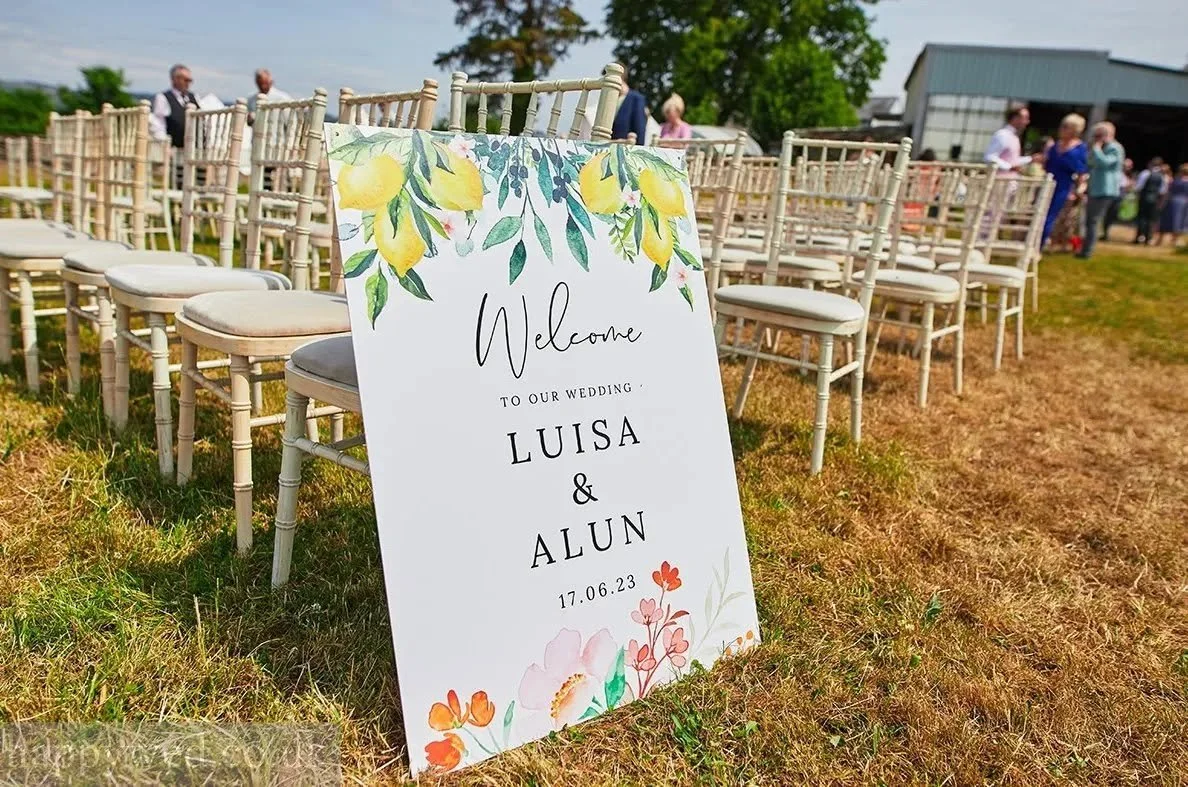 Wedding welcome sign with floral design and names Luisa and Alun, date 17.06.23, placed outdoors near rows of chairs and guests.