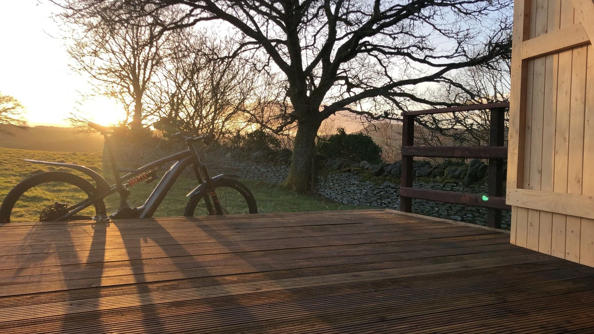 Sunset over a rural landscape with a bare tree, a stone wall, and a bicycle leaning against a wooden deck.