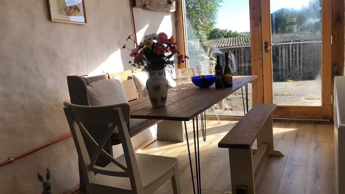 A wooden dining table with a vase of flowers, two bottles, and a bowl sits near a glass door leading outside to a paved yard. A white chair and a bench are positioned at the table, and sunlight brightens the room.