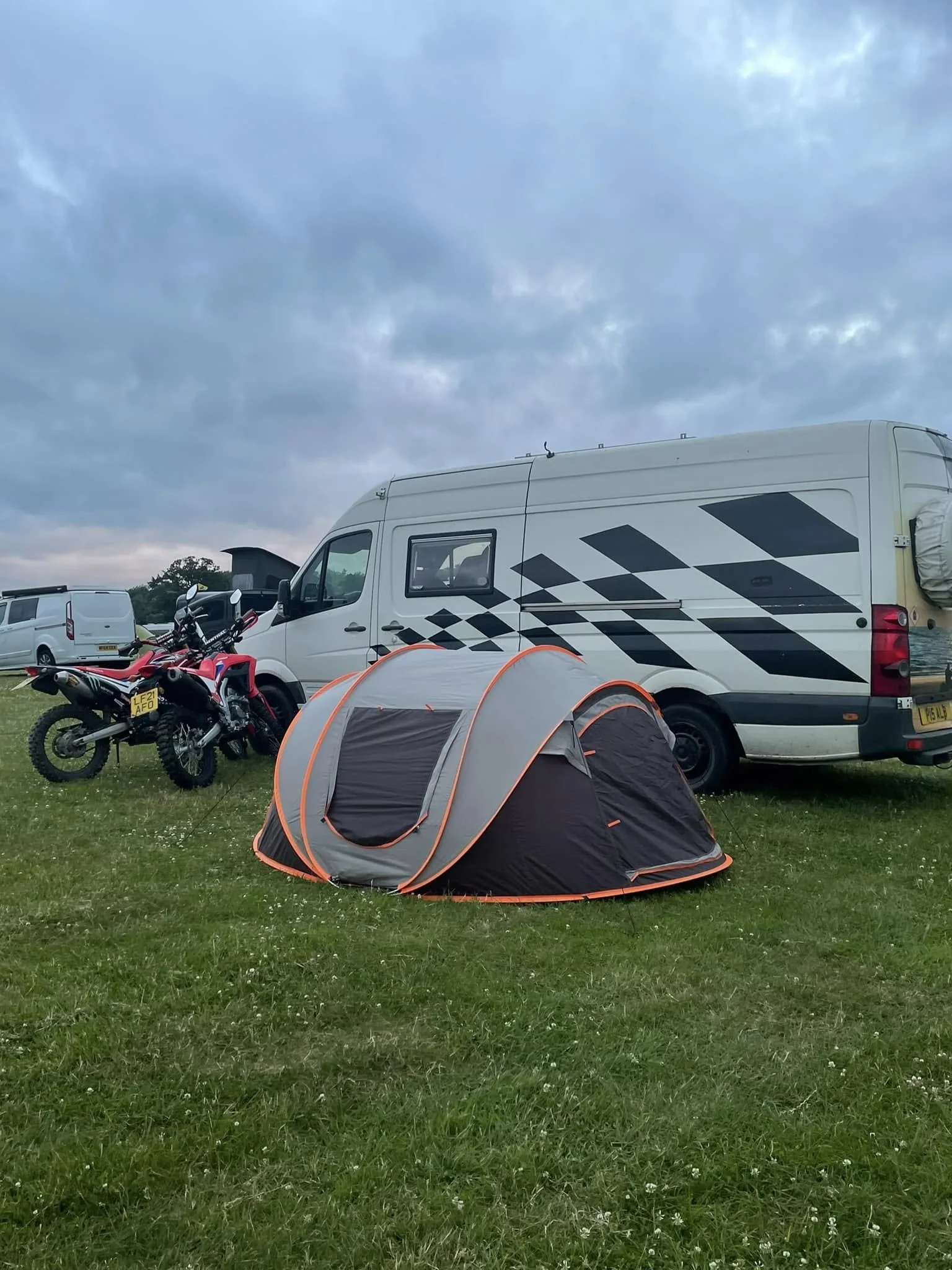 Camping scene with a gray and orange tent on green grass, a black and red motorcycle, a white camper van, and a cloudy sky.