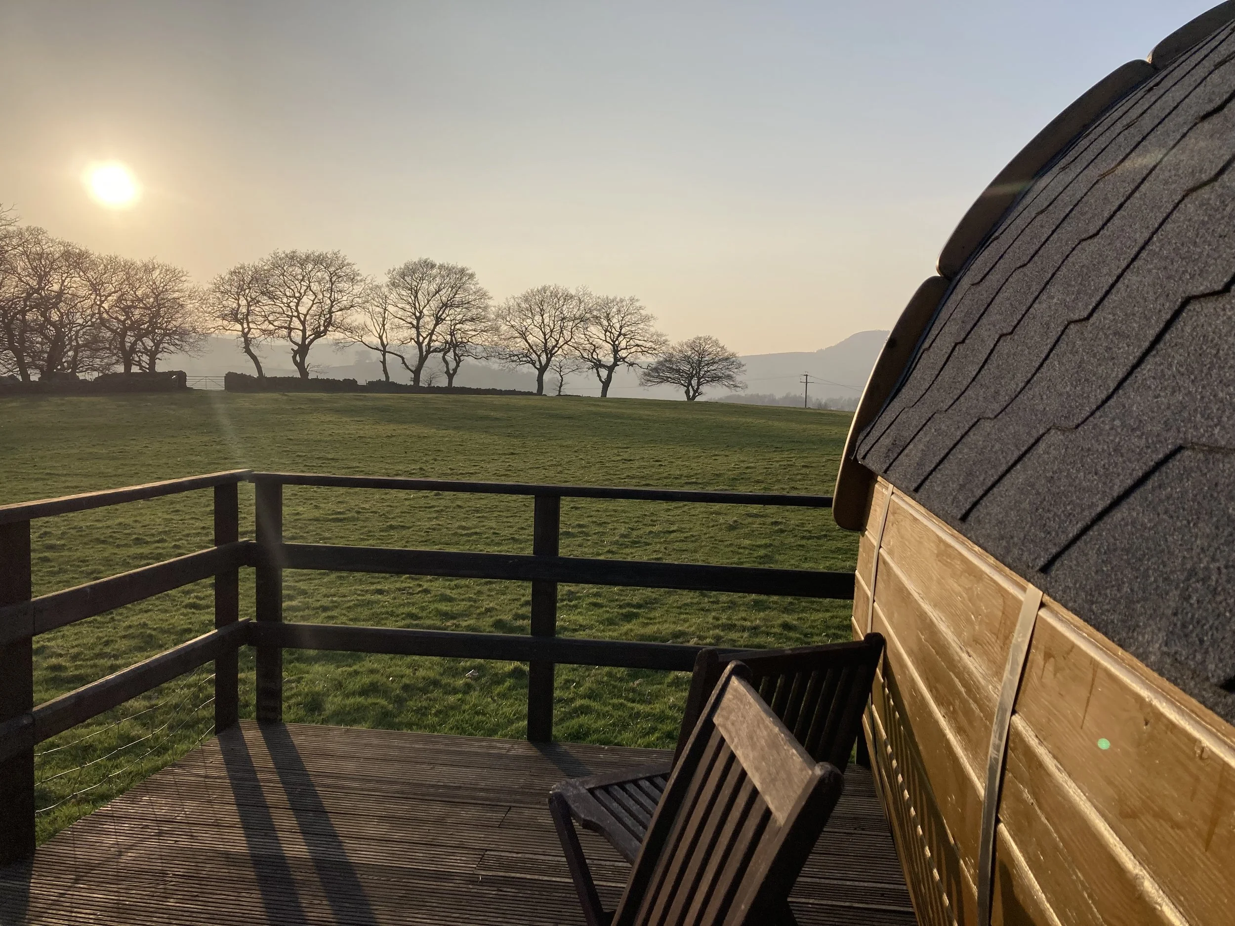 Sunset over a field with leafless trees in the distance, viewed from a wooden deck that has benches and a railing, with part of a building's roof visible on the right.